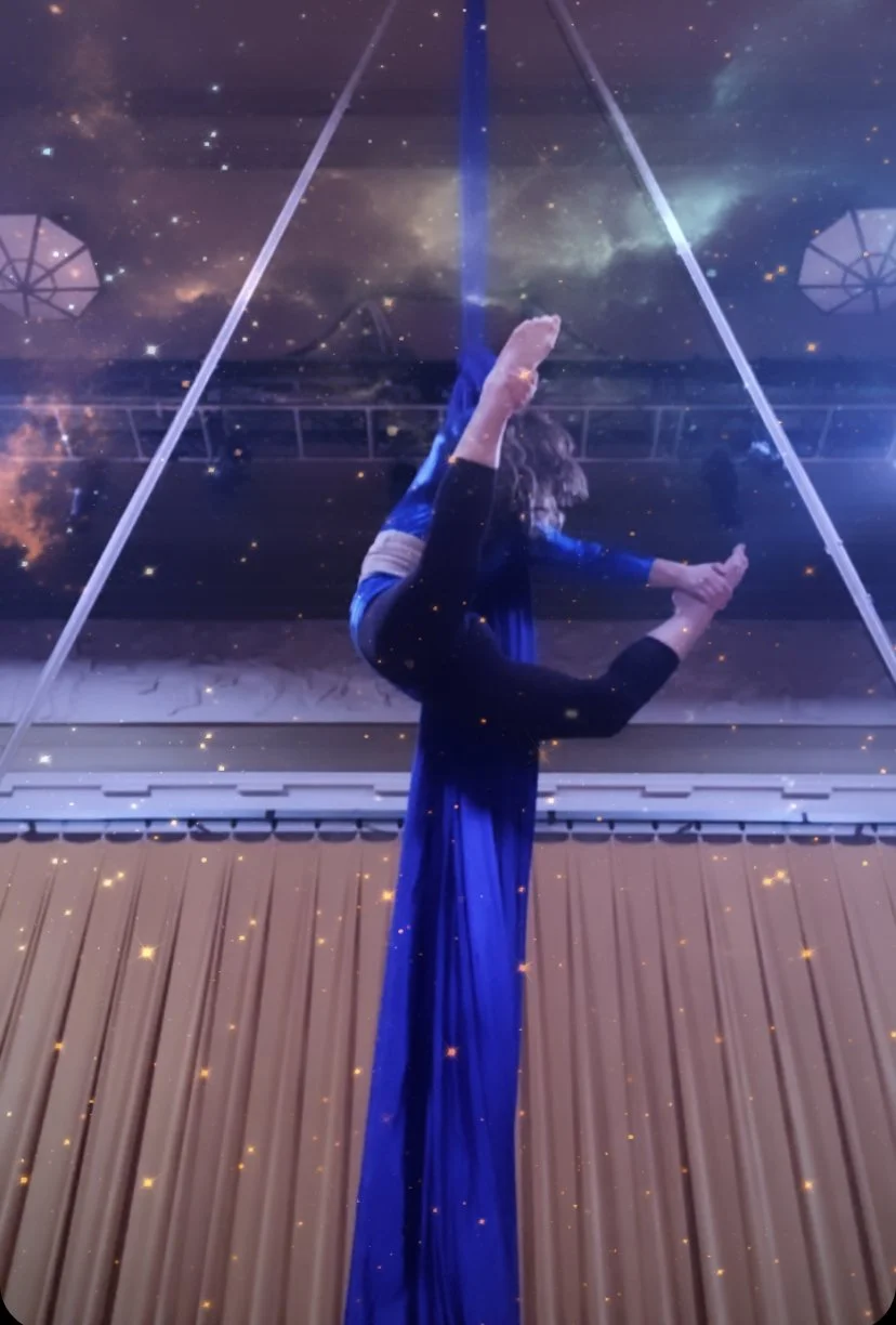 A person performing an aerial yoga pose on a fabric hammock with a starry galaxy and nebula background, viewed from below through a glass ceiling.