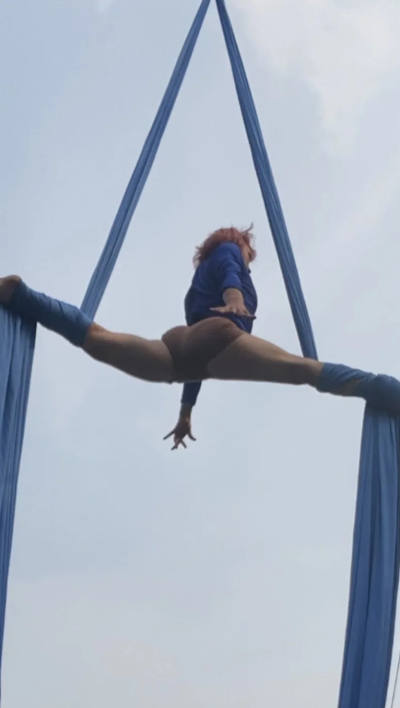 A person with red hair in a blue jacket performing an aerial split on blue aerial silks outdoors against a cloudy sky.
