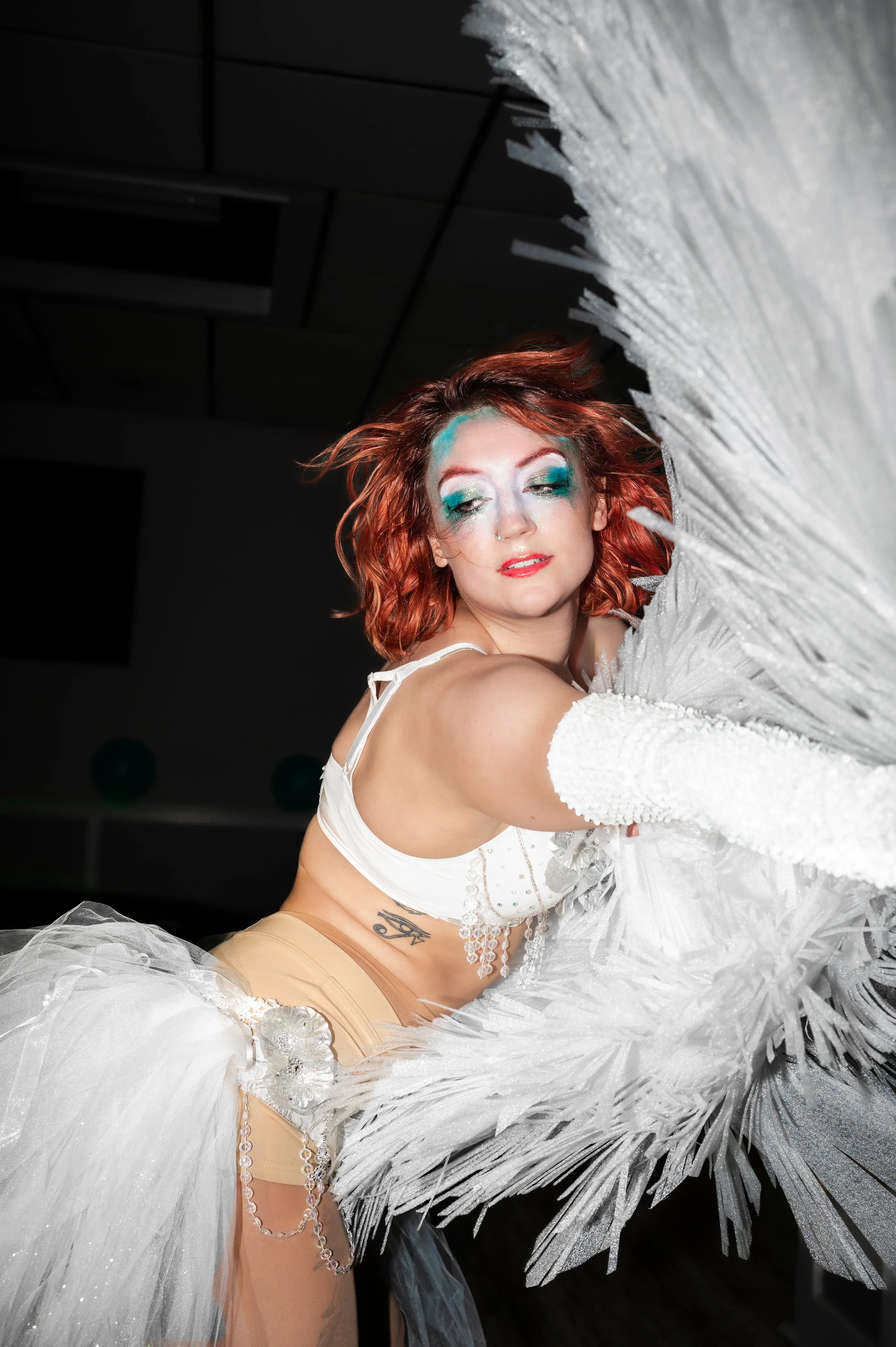 A woman with red hair and dramatic makeup, wearing a white top and beige shorts, posing with her eyes closed amidst large white feathers.