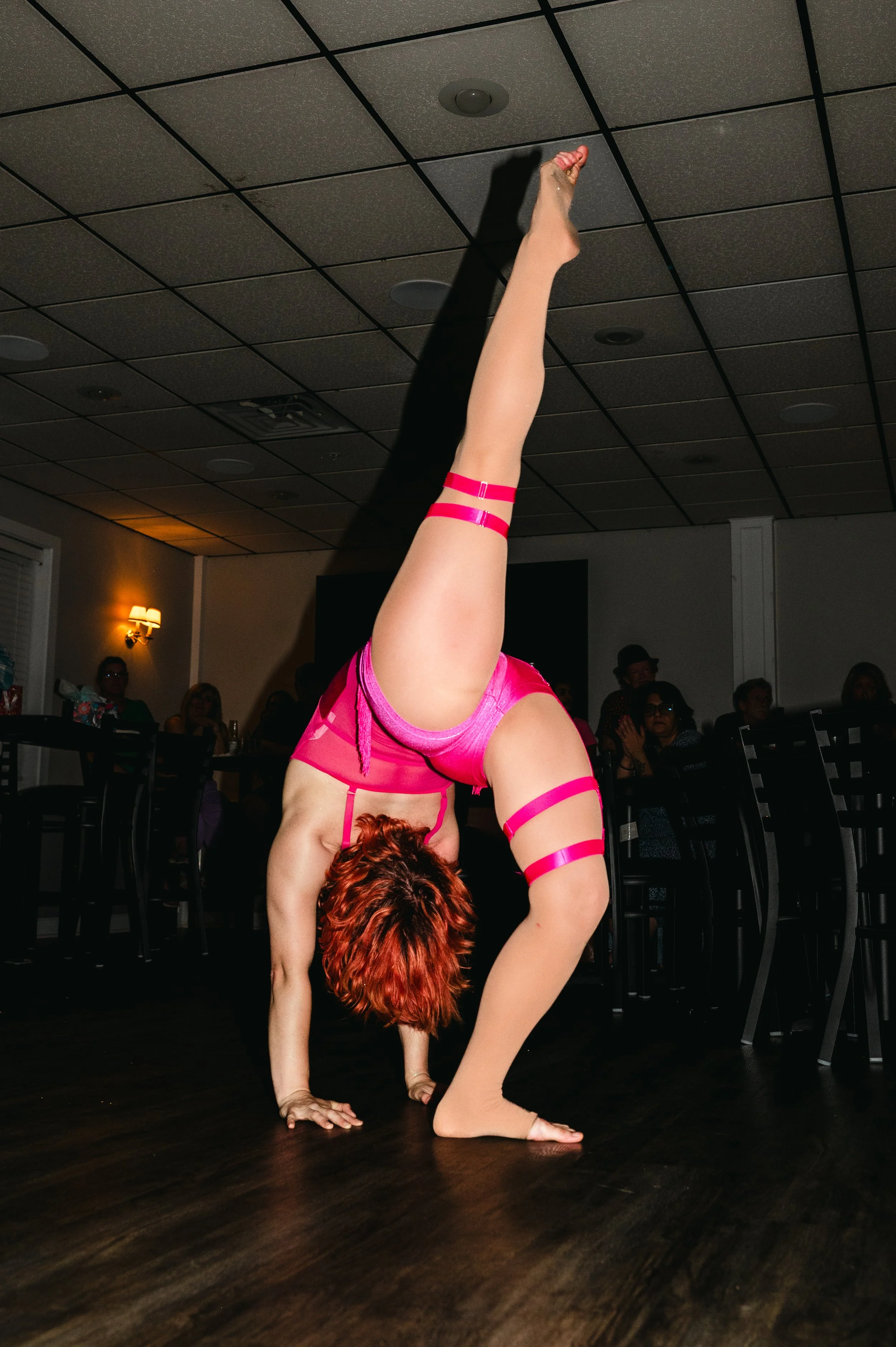 A performer in a pink costume doing a handstand on a dark wooden floor during an indoor event, with an audience watching in the background.