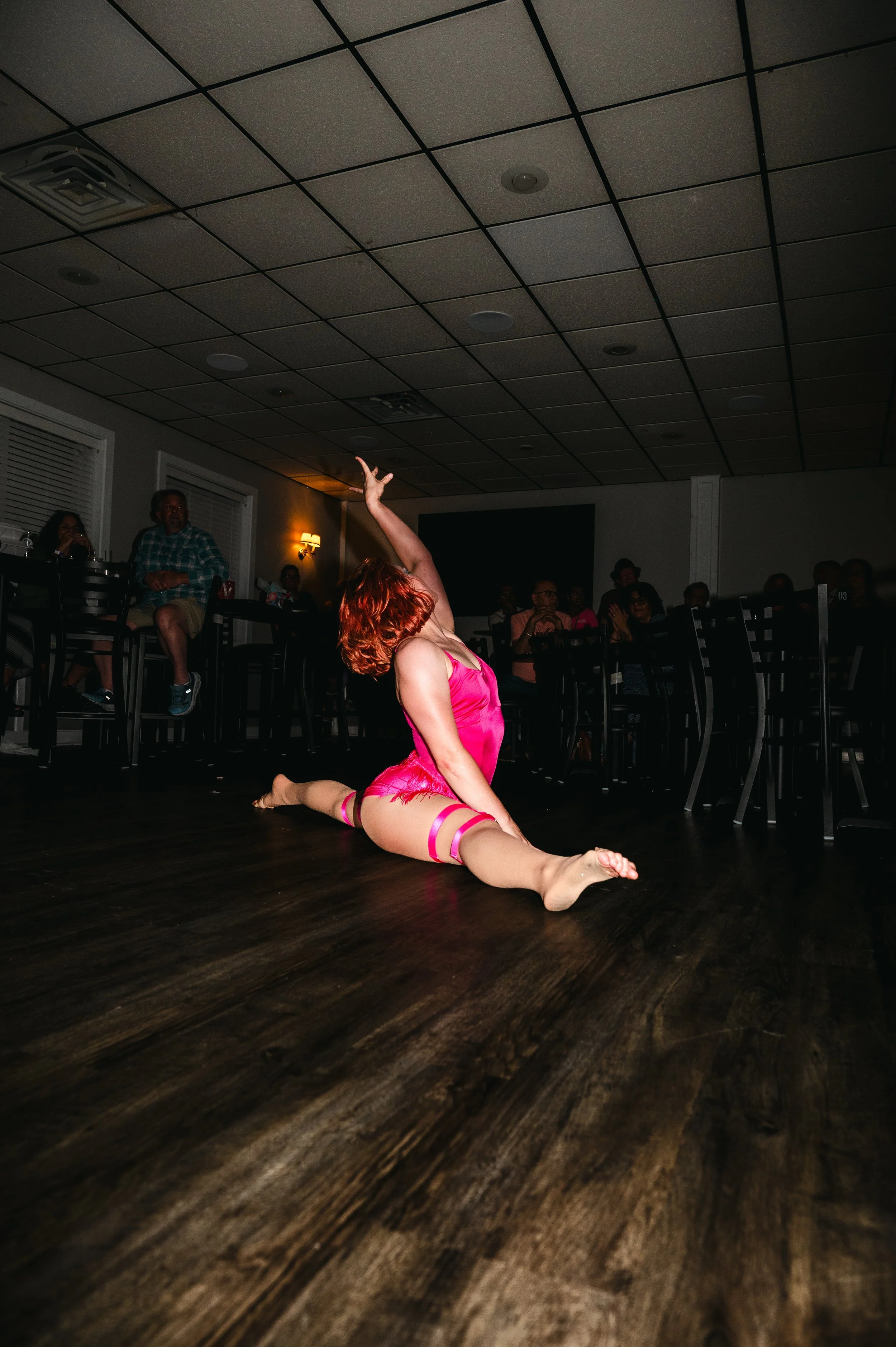 A woman with red hair in a pink dance costume performing a split on a wooden dance floor during a performance. She is raising one arm above her head, and an audience seated at tables watches in the background.