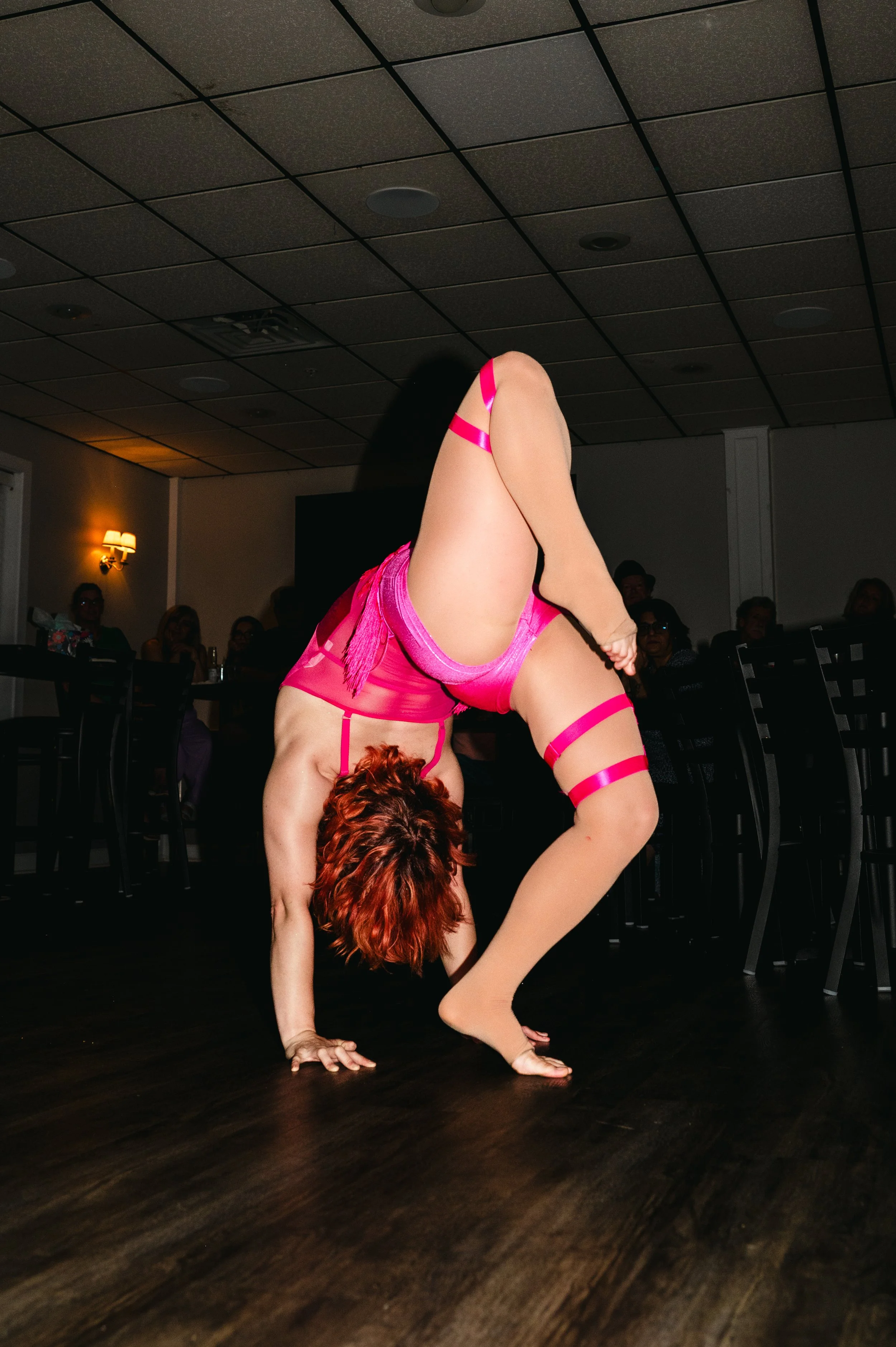 A performer in a pink costume is in an upside-down handstand pose on a wooden floor with audience members seated in the background.