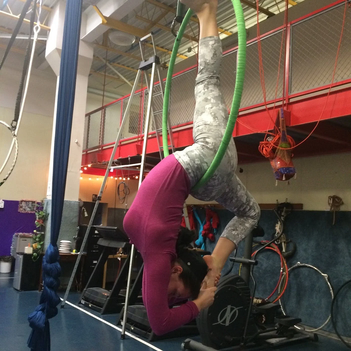 A woman performing an aerial acrobatics pose on green aerial hoops, with one leg extended upward and the other bent, while holding hands with a person lying on the ground. The setting appears to be an indoor trampoline or aerial arts gym.