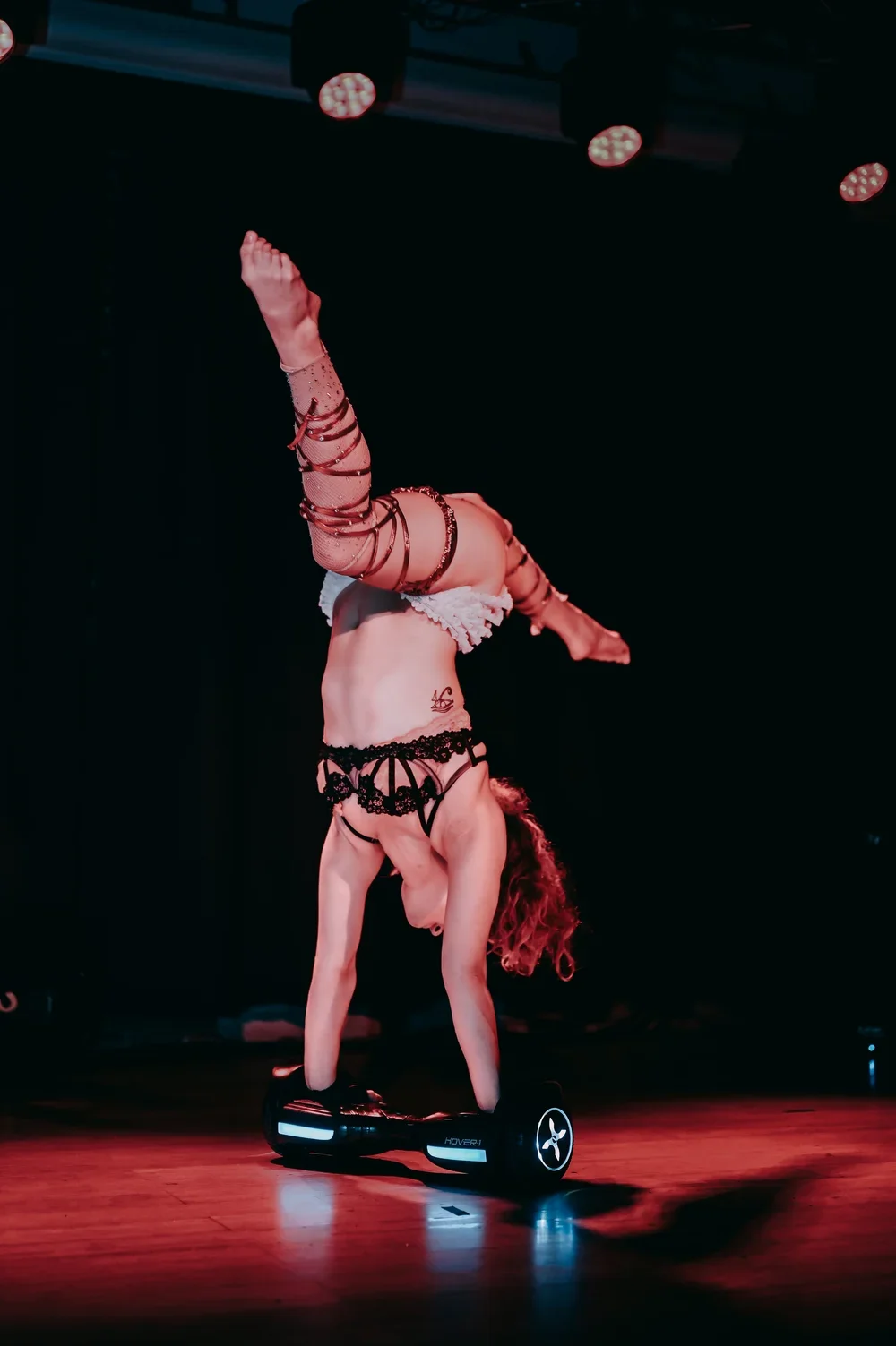 Talia Hazard, seen balancing upside down on a hoverboard with her hands on the floor, wearing a black lingerie set, lace arm warmers, and stockings, on a stage with red lighting.
