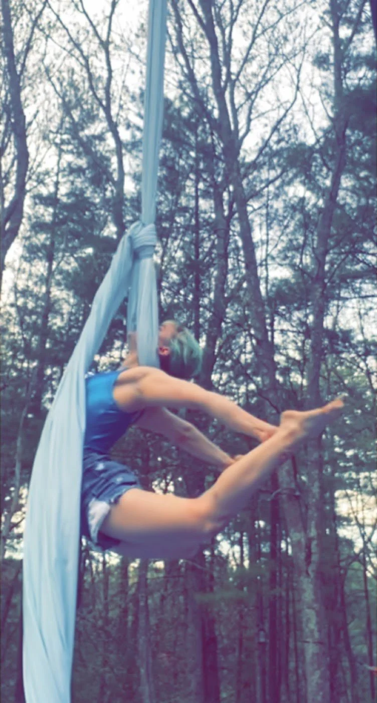 A woman with dyed green hair is performing aerial silk acrobatics outdoors among tall trees, hanging upside down with her legs wrapped around the silk and arms extended forward.