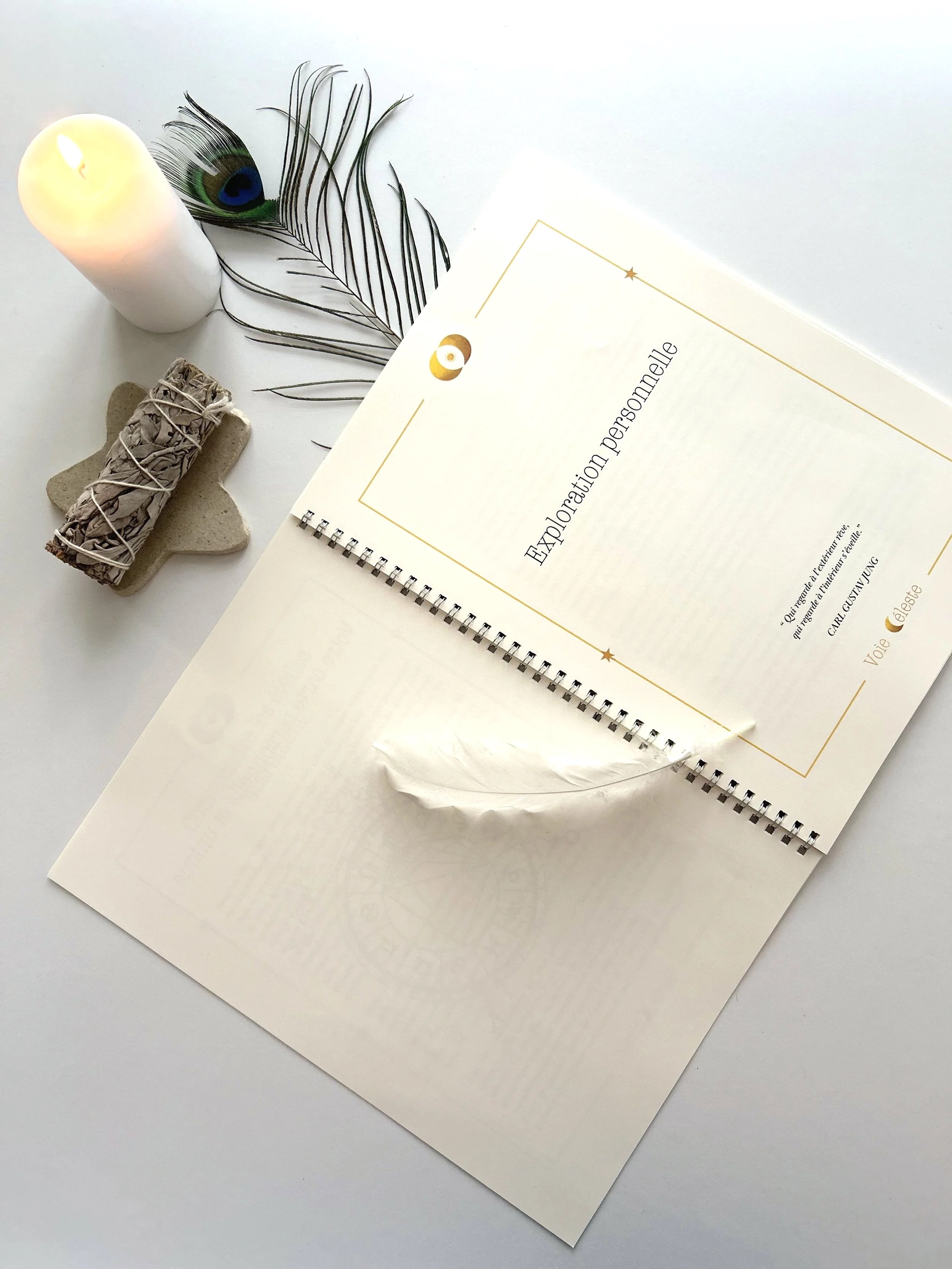 Une brochure intitulée "Exposition personnelle" à côté d'une plume de paon, d'une bougie et d'une petite pierre avec un cylindre en saule tortueux.