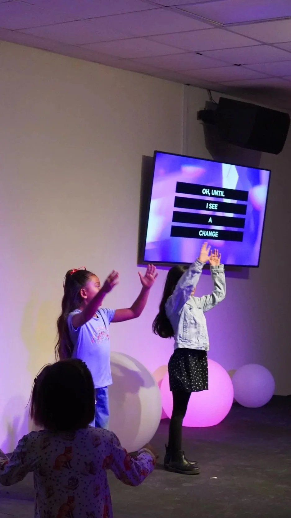 Children singing on stage with a large screen displaying the lyrics 'Oh, until I see a change' in the background.