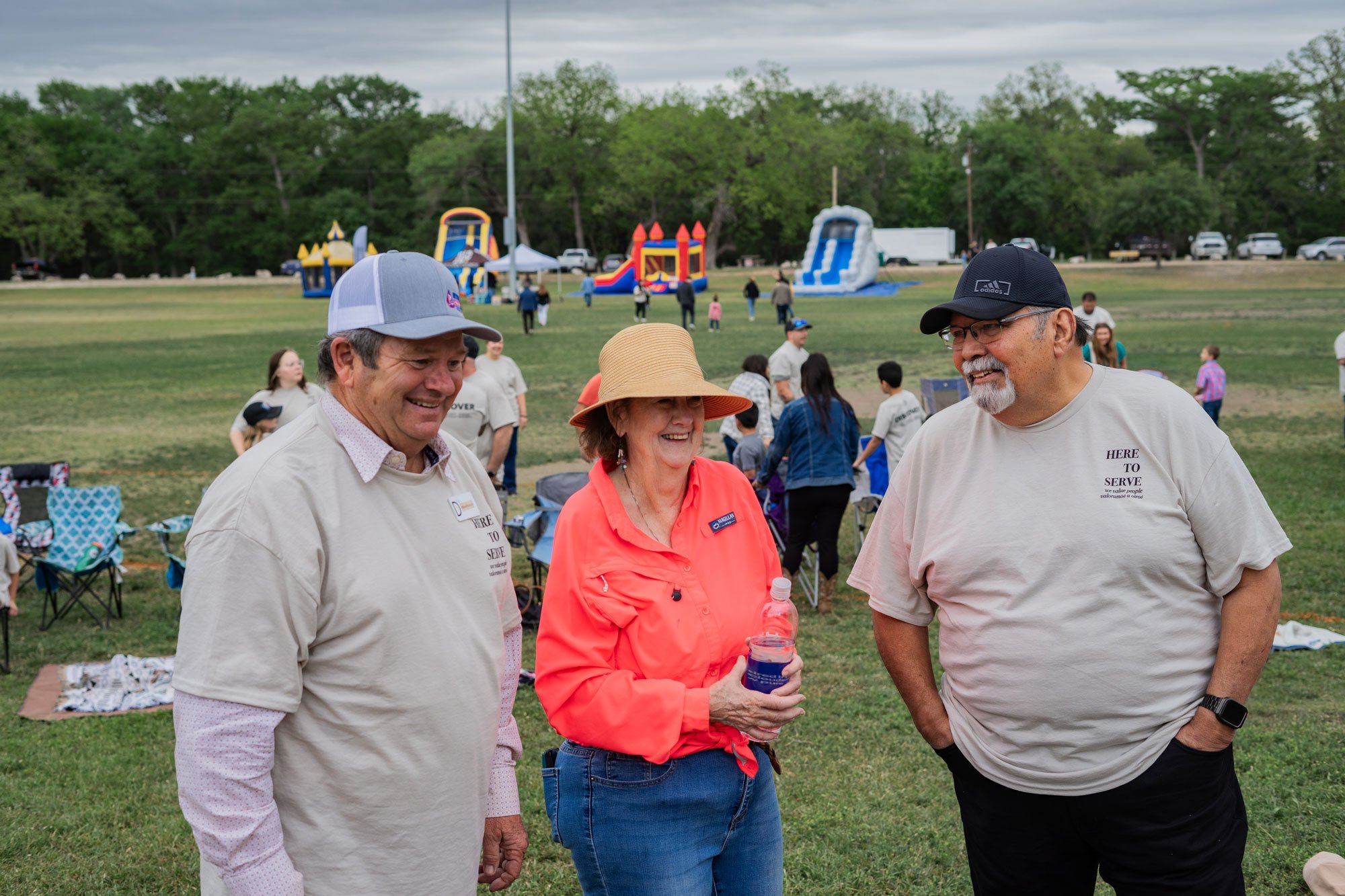 Three smiling people talking in a park during an outdoor event, with inflatable slides and picnic chairs in the background.