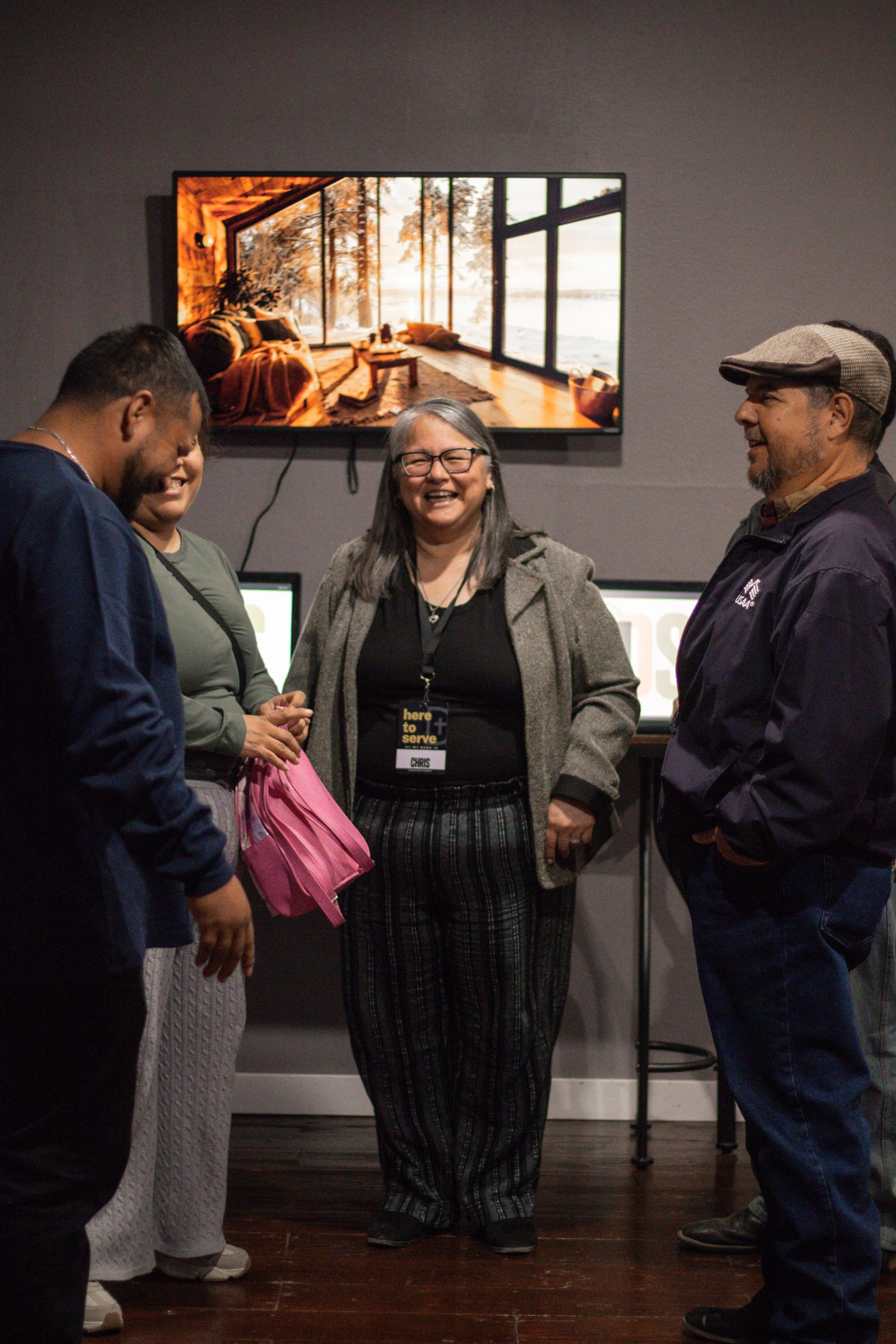 A group of four people standing and talking in an indoor setting, with a woman in the center laughing. Behind them is a large screen showing a cozy room with a large window overlooking a scenic landscape.