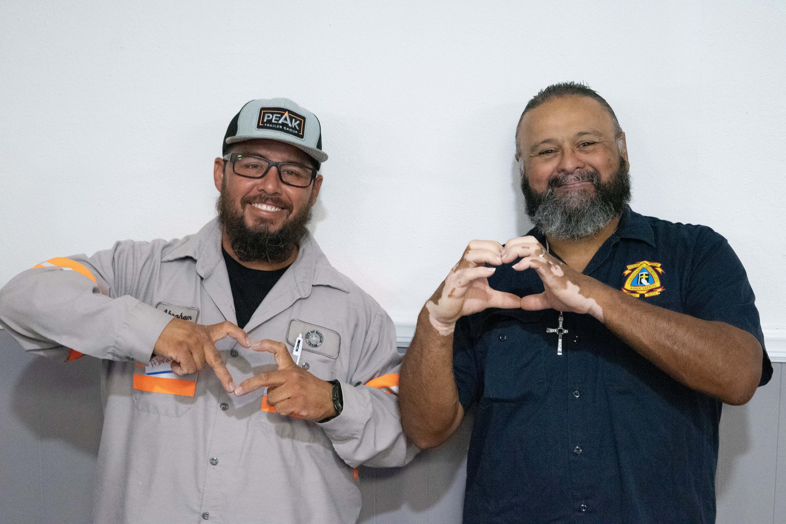 Two men standing side by side making heart shapes with their hands. The man on the left is wearing glasses, a gray work uniform with orange accents, and a trucker hat. The man on the right is wearing a dark blue shirt with a badge on it. Both are smiling.