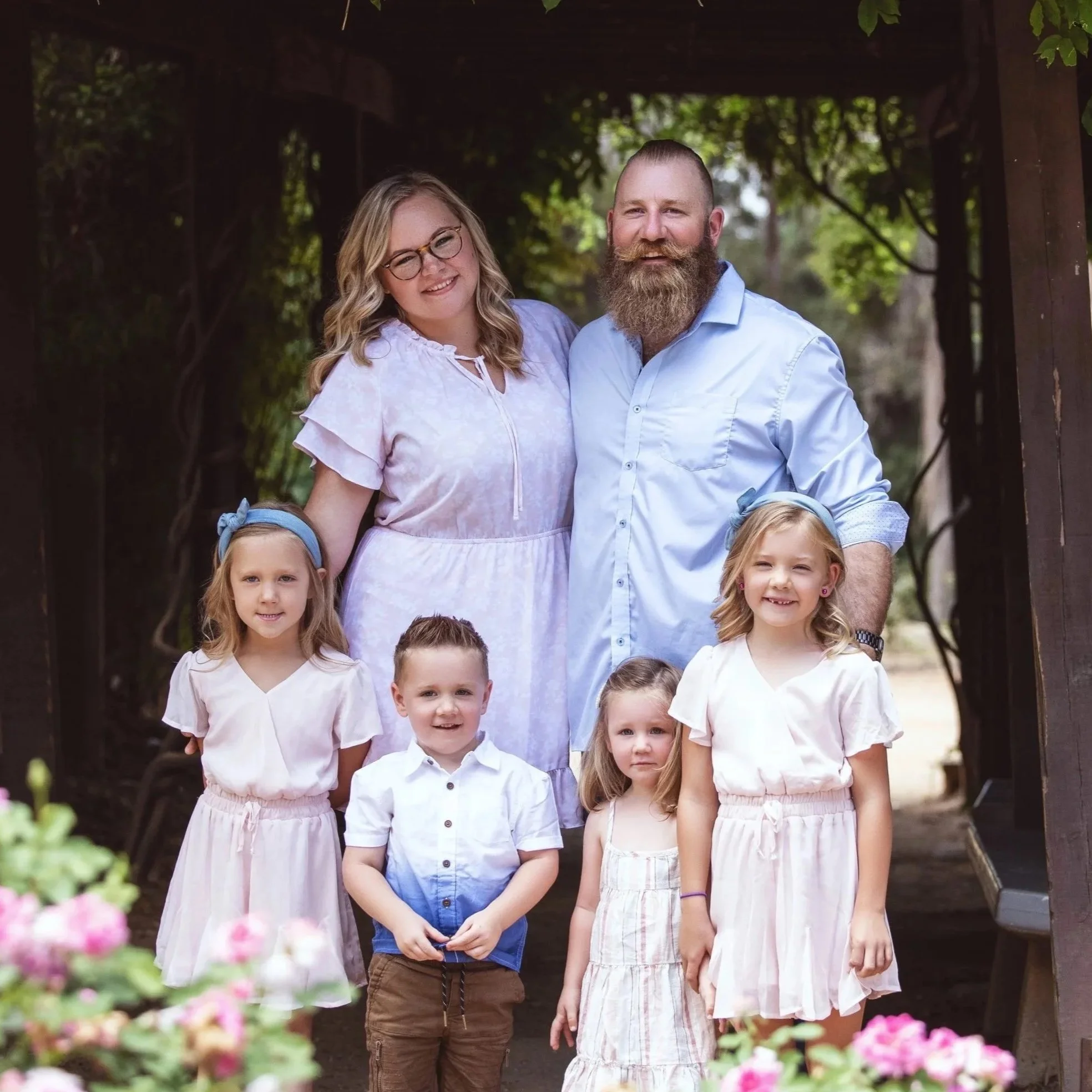 Family of six, including father, mother, and four children, standing together outdoors under a wooden arch, surrounded by trees and pink flowers.