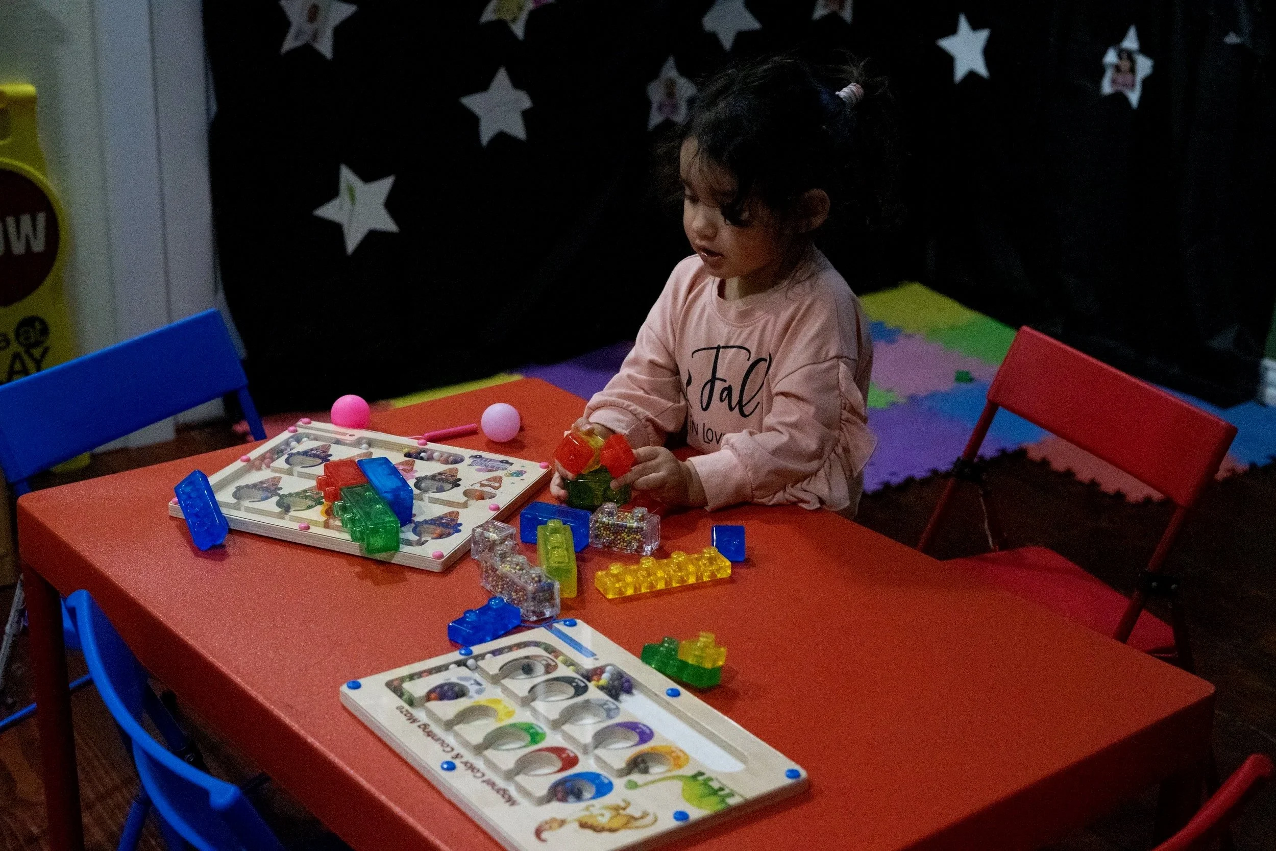 A young girl with dark curly hair tied with beads sits at a red table playing with colorful building blocks and game pieces. Two blue chairs are positioned on the left, and a red folding chair is on the right. The background features a black curtain with star shapes and a rainbow-colored foam mat on the floor.