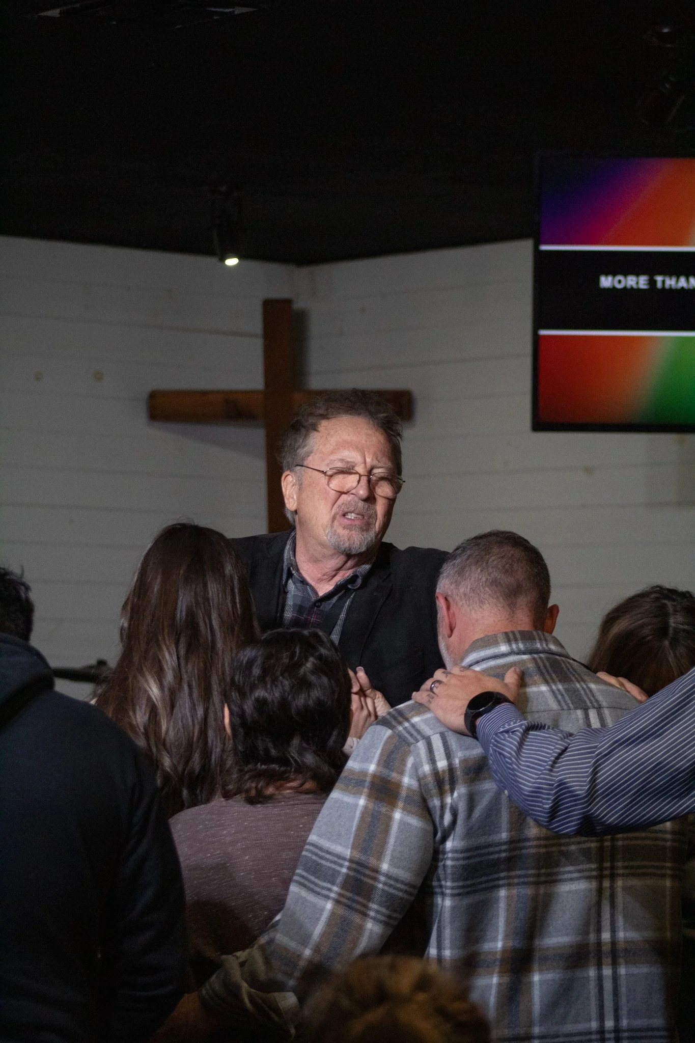 A man with glasses and a beard is praying with a group of people inside a church, with a cross on the wall behind him.