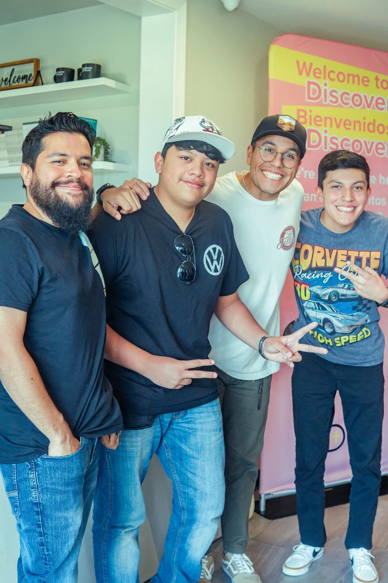 Four men standing together indoors, smiling, with a pink banner behind them that reads 'Welcome to Discovery' in multiple languages.