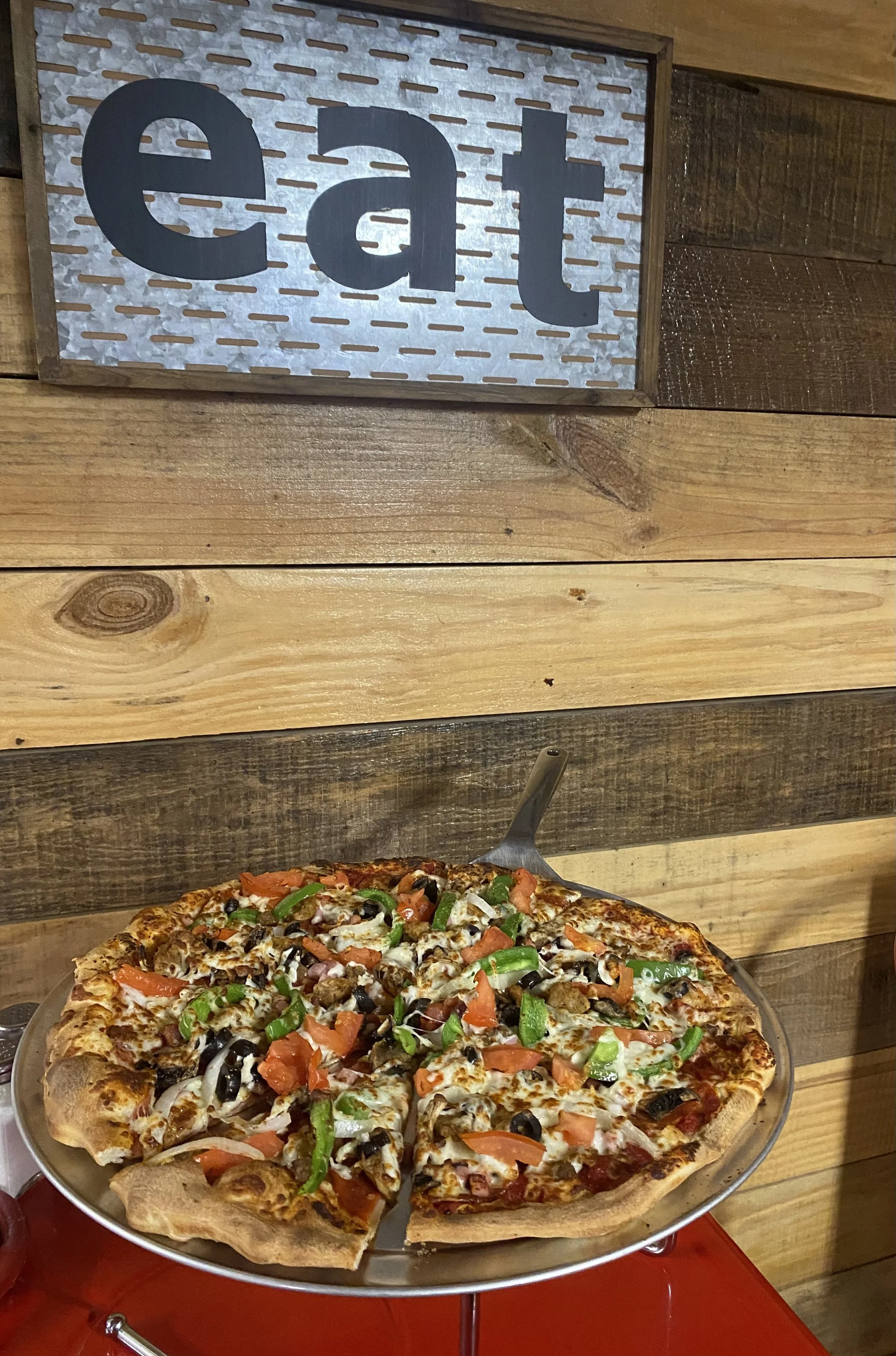 A pepperoni, green pepper, black olive, and mushroom pizza on a silver tray in front of a wooden wall with an 'eat' sign.