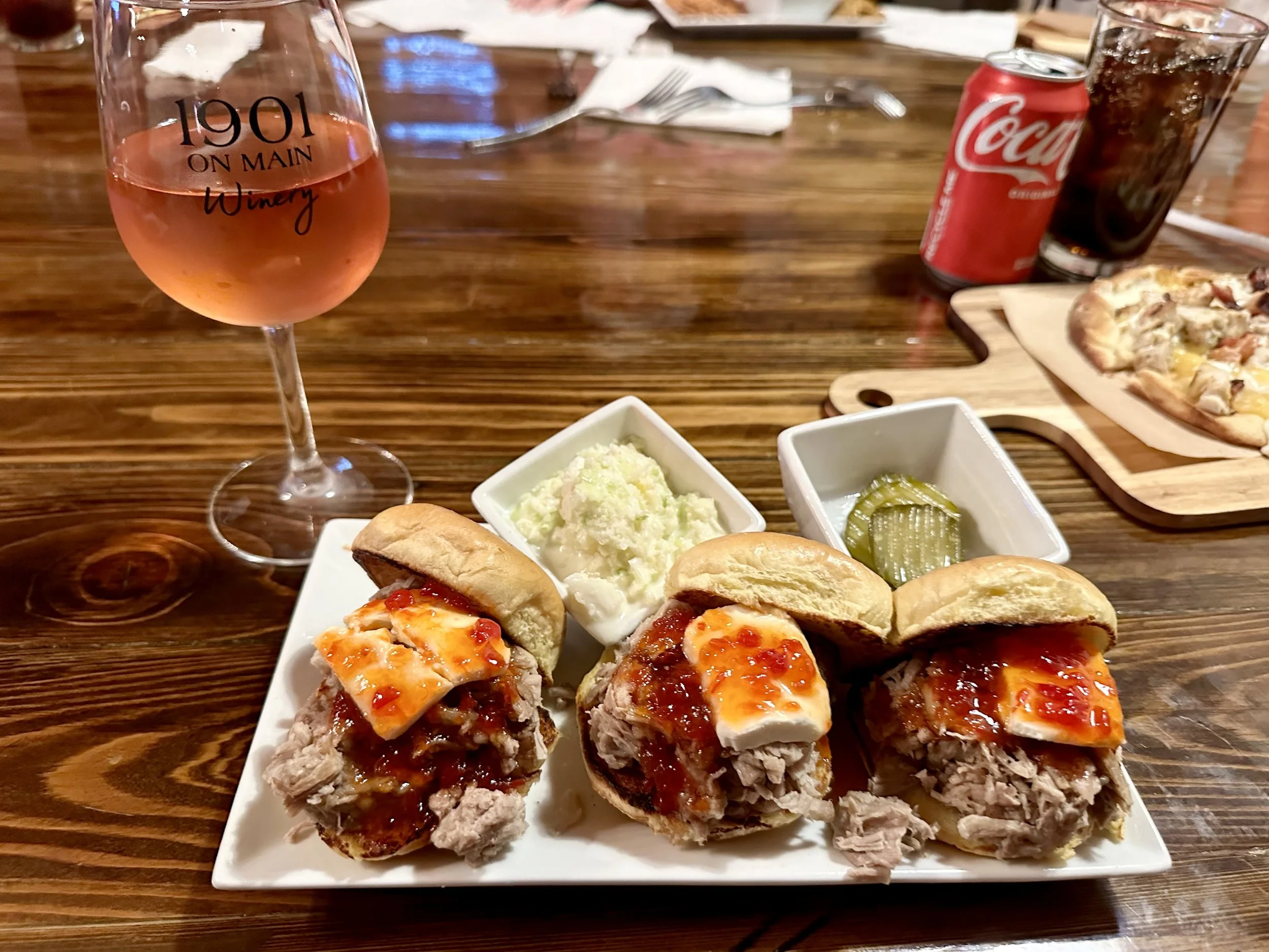 A plate of three barbecue pork sandwiches topped with sauce and cheese, served with two side dishes of potato salad and pickles, on a wooden table with a glass of rosé wine, a can of Coca-Cola, a glass of cola, and a pizza in the background.