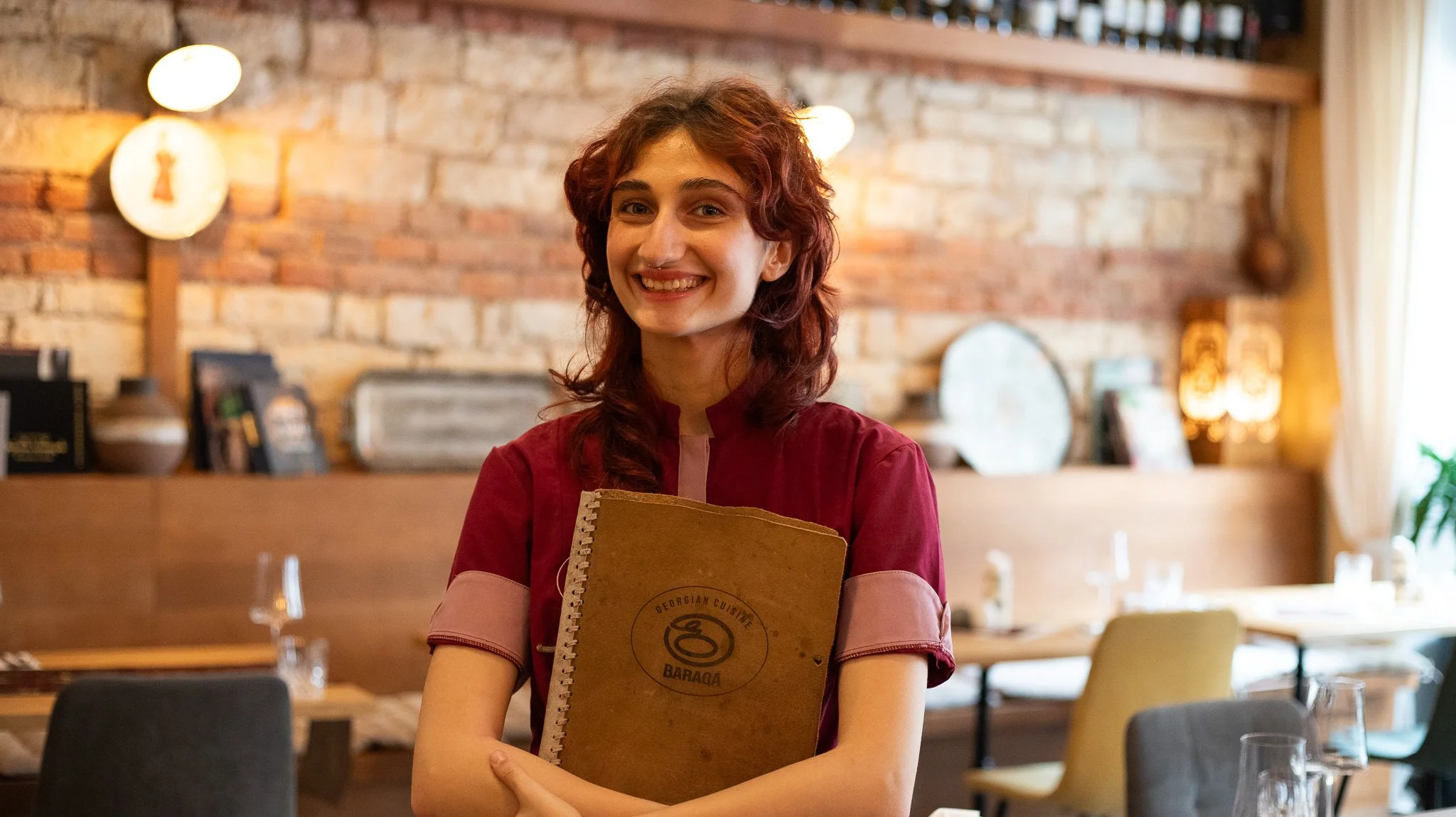A smiling woman with red hair holding a brown menu in a warmly lit restaurant with a rustic brick wall background.