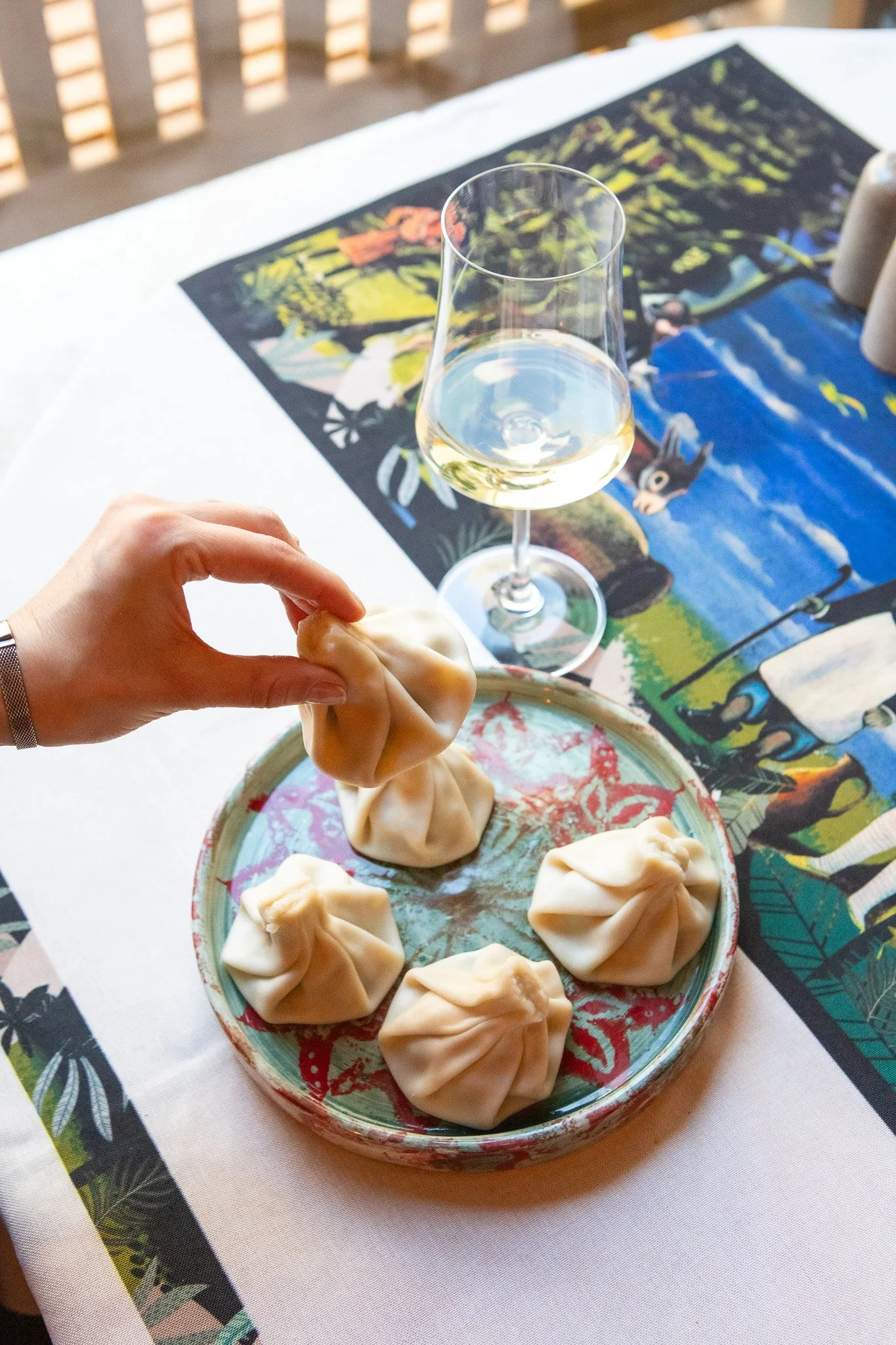 A hand picking up a dumpling from a plate with five dumplings, a glass of white wine, and a colorful tablecloth with illustrated outdoor scene in the background.