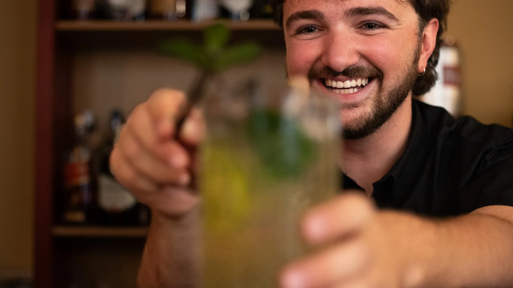 A man with a beard and earring smiling and holding a glass with a cocktail and mint leaves, in a bar or cafe setting.