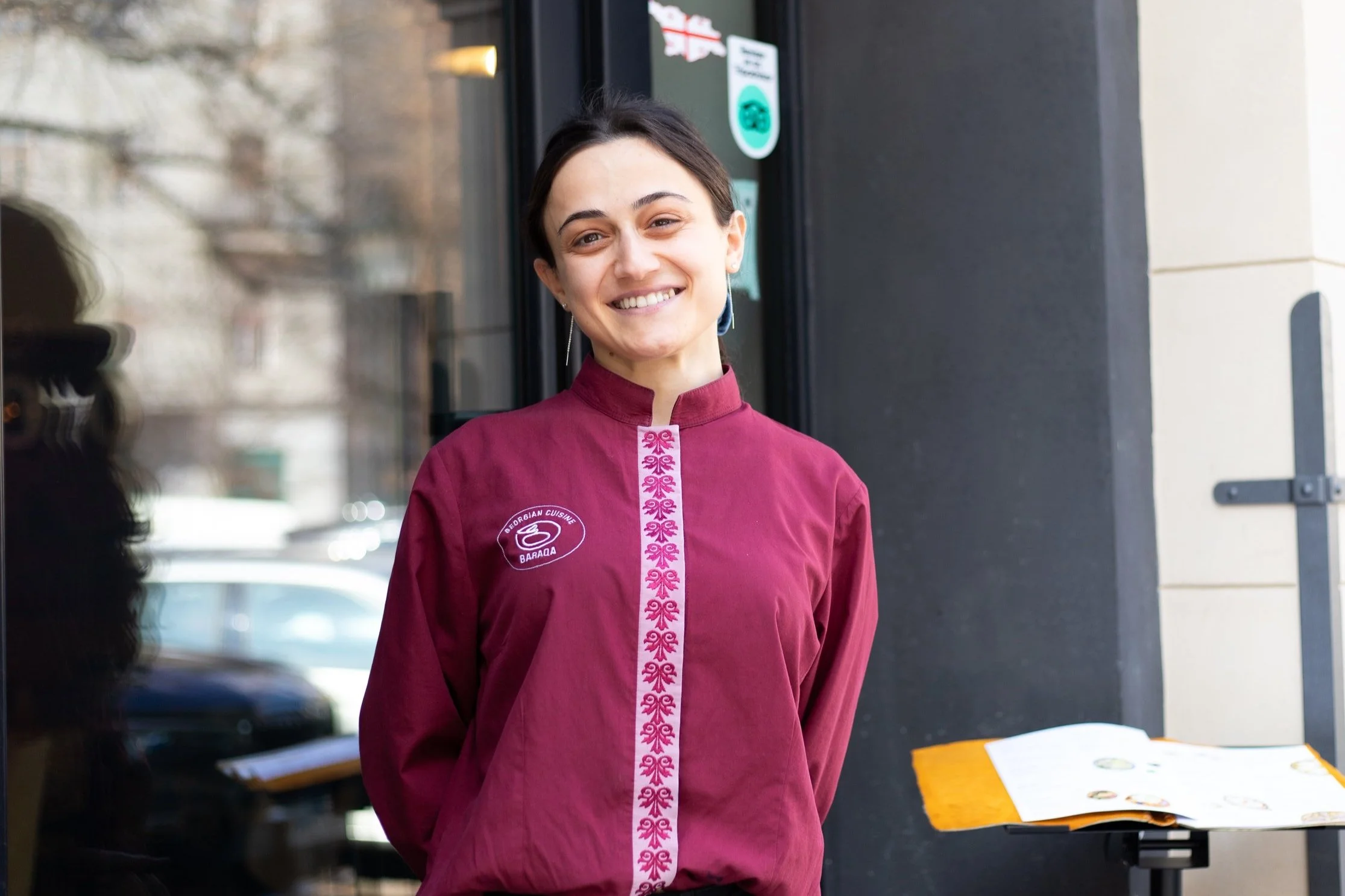 A woman wearing a maroon chef jacket with traditional embroidery standing outside a restaurant.