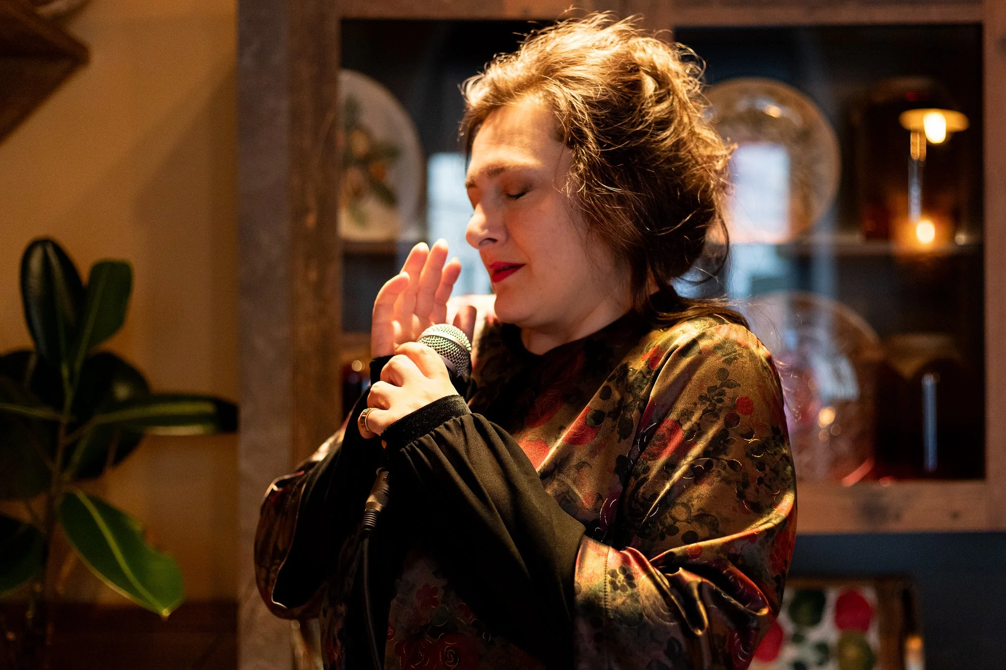 A woman with curly hair, closed eyes, and red lipstick is holding a microphone and appears to be singing or speaking with her hand near her face in a cozy, warmly lit room.