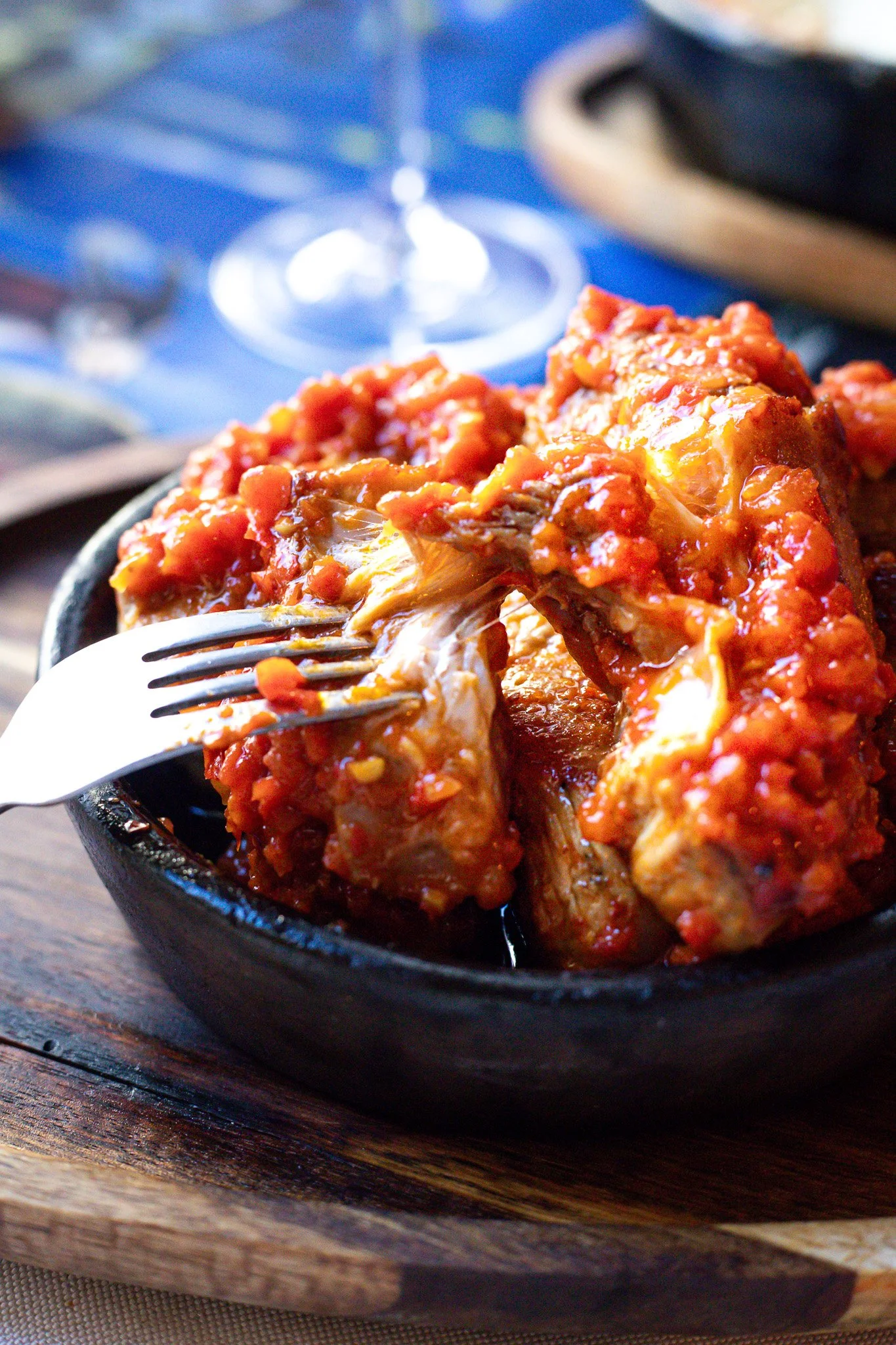Close-up of a bowl of chicken wings covered in spicy red sauce on a wooden tray, with a fork piercing into one wing, set on a table with a blue tablecloth and a glass of wine in the background.