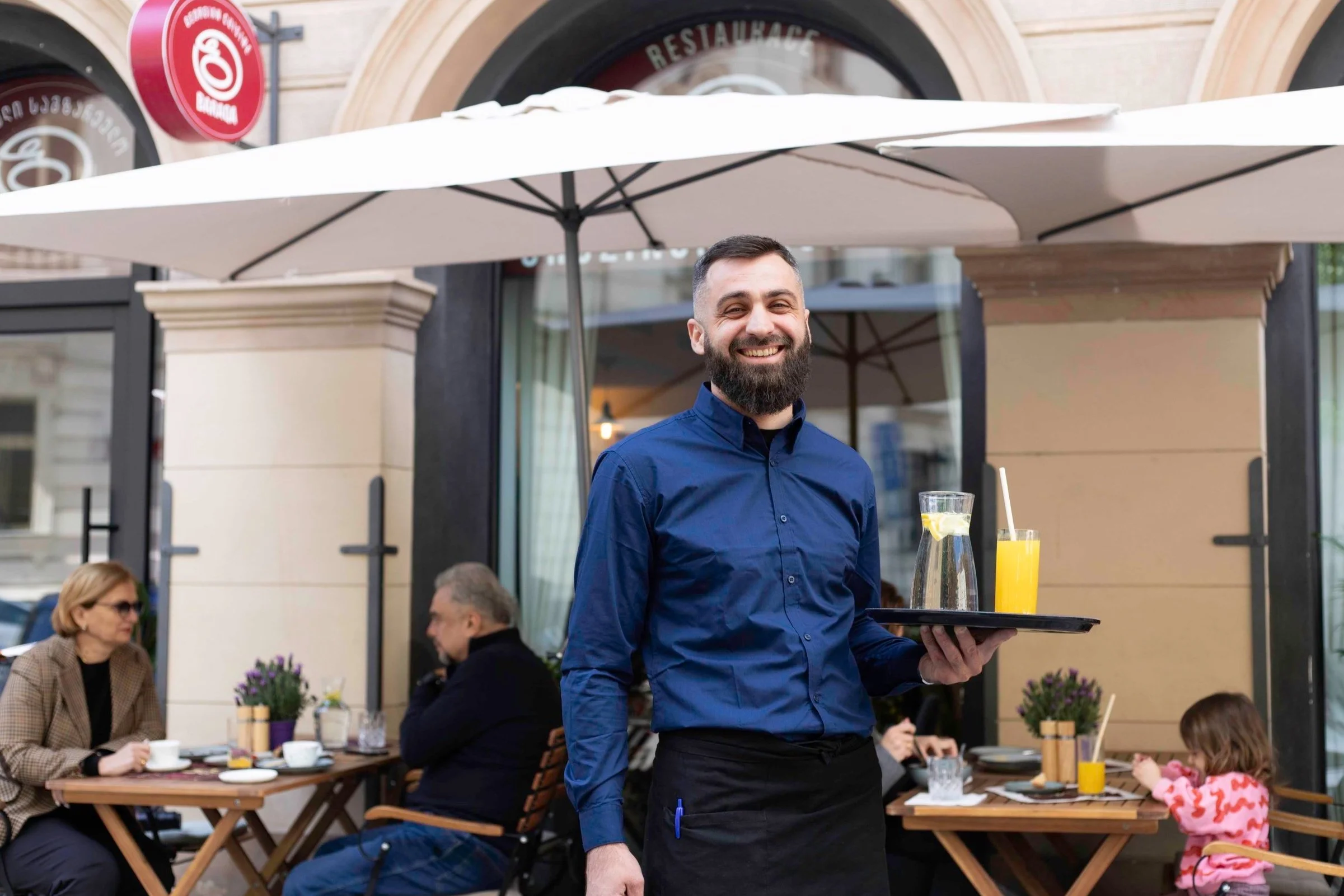 A smiling waiter in a blue shirt and black apron holding a tray with a pitcher of water and a glass of orange juice at an outdoor restaurant patio.