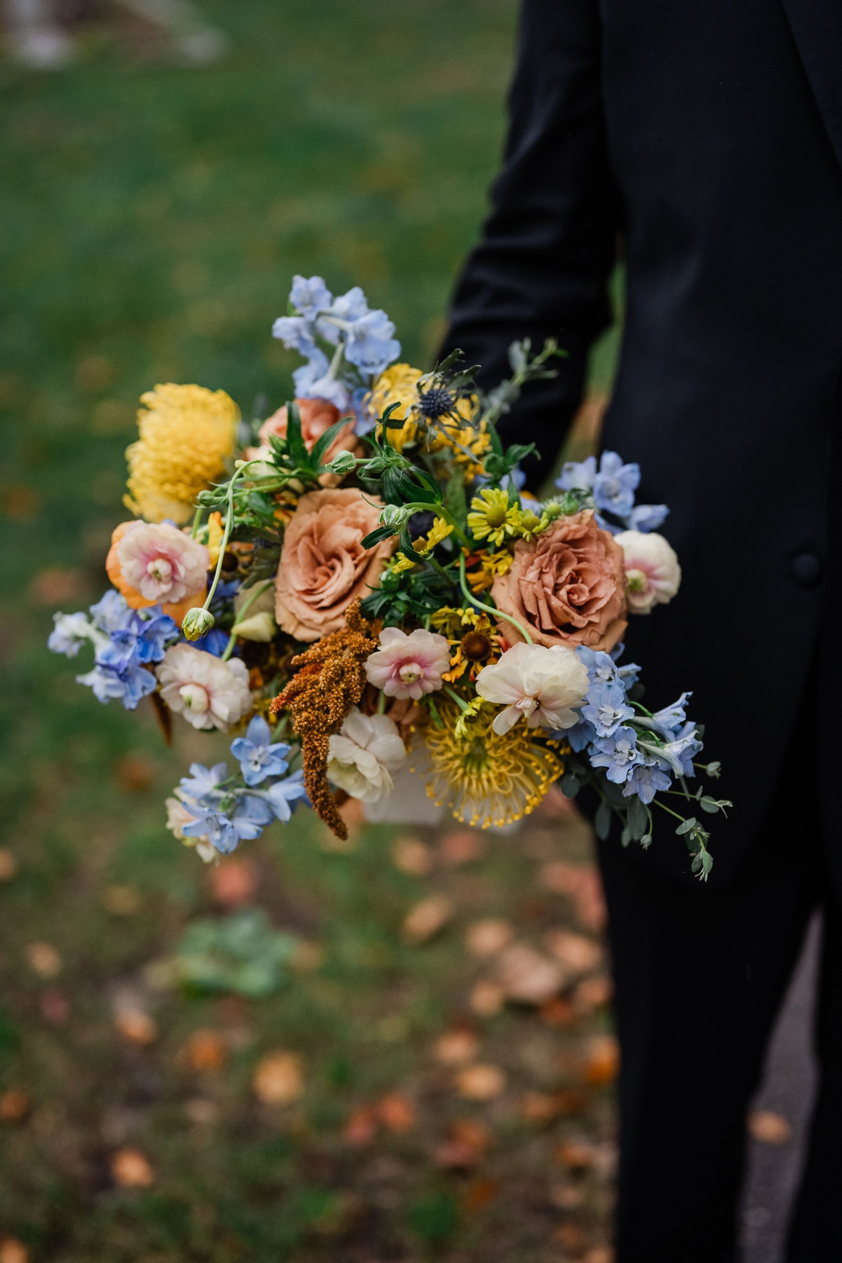 Person in a black suit holding a colorful bouquet of flowers outdoors.