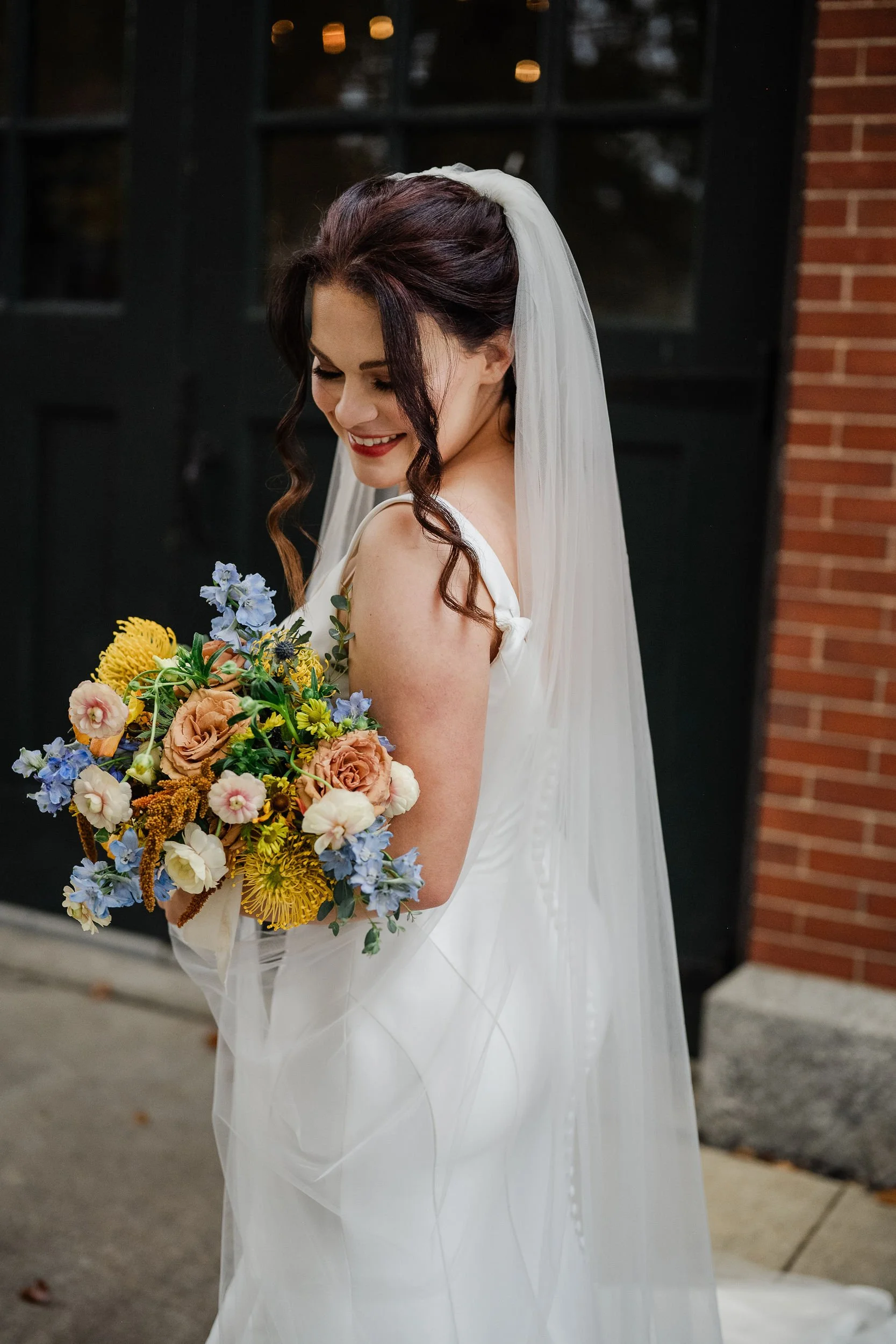 Smiling bride holding a colorful bouquet of flowers outdoors, wearing a white wedding dress and veil, standing near a brick and black door, with her hair styled in loose curls.