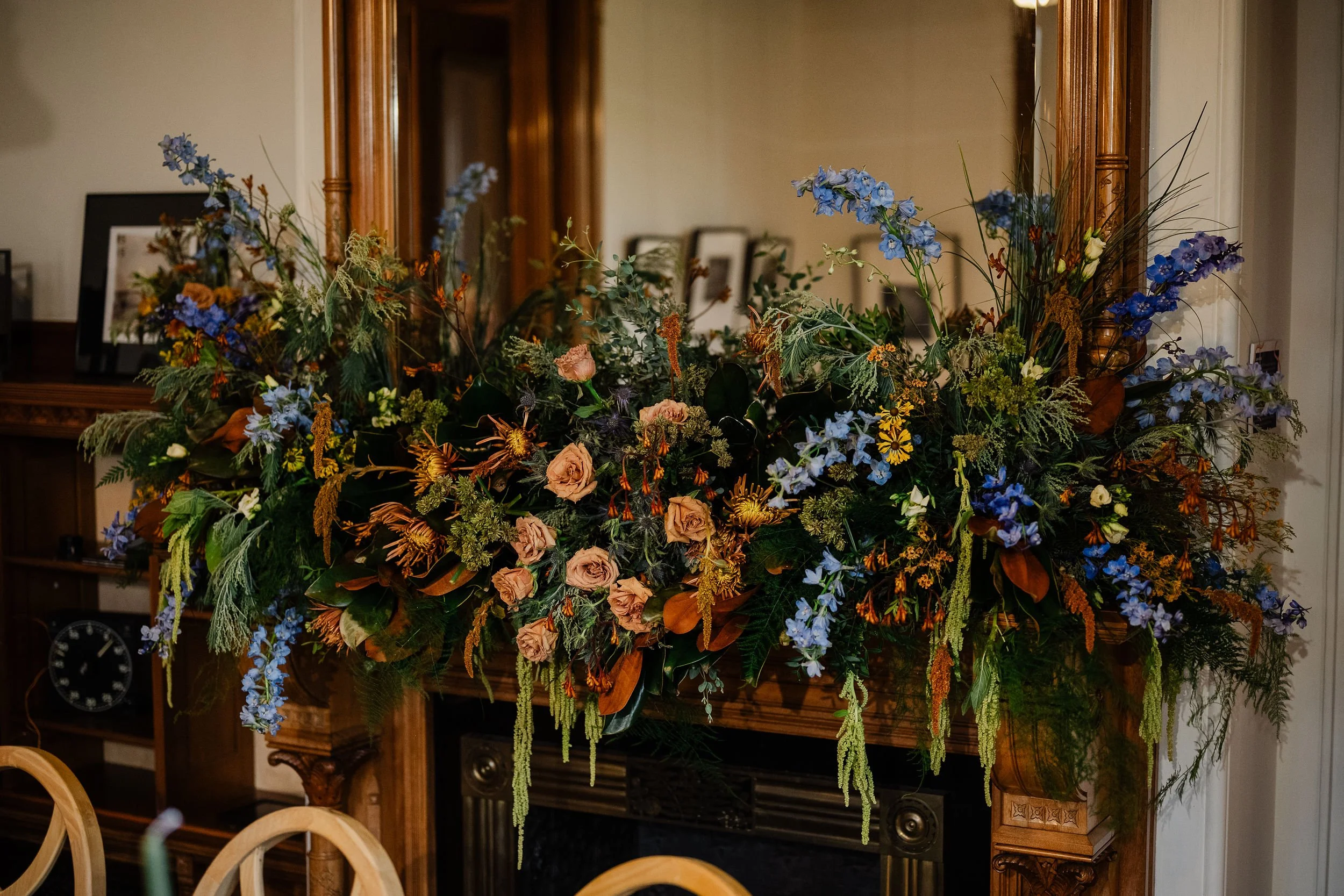 A large floral arrangement with blue, purple, and peach-colored flowers on a wooden fireplace mantle in a room with framed photos and a mirror.