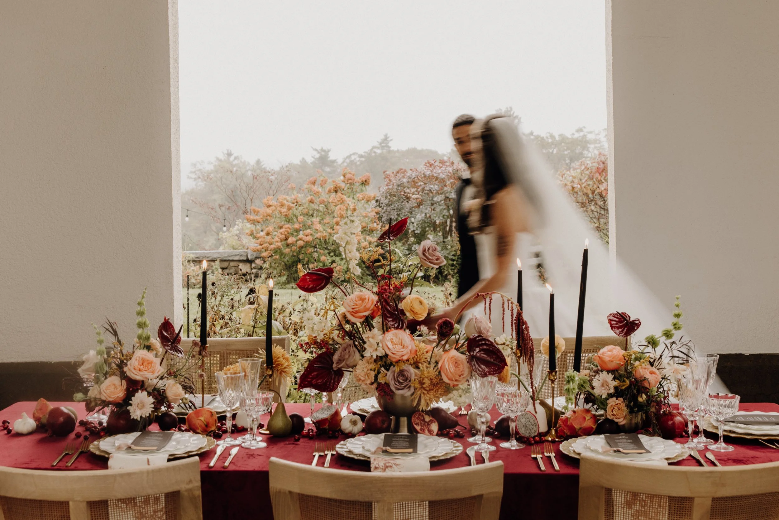 A festive dining table decorated with pink and red flowers, black candles, crystal glassware, and assorted fruits, set in front of a large window with a view of blooming trees during daytime.