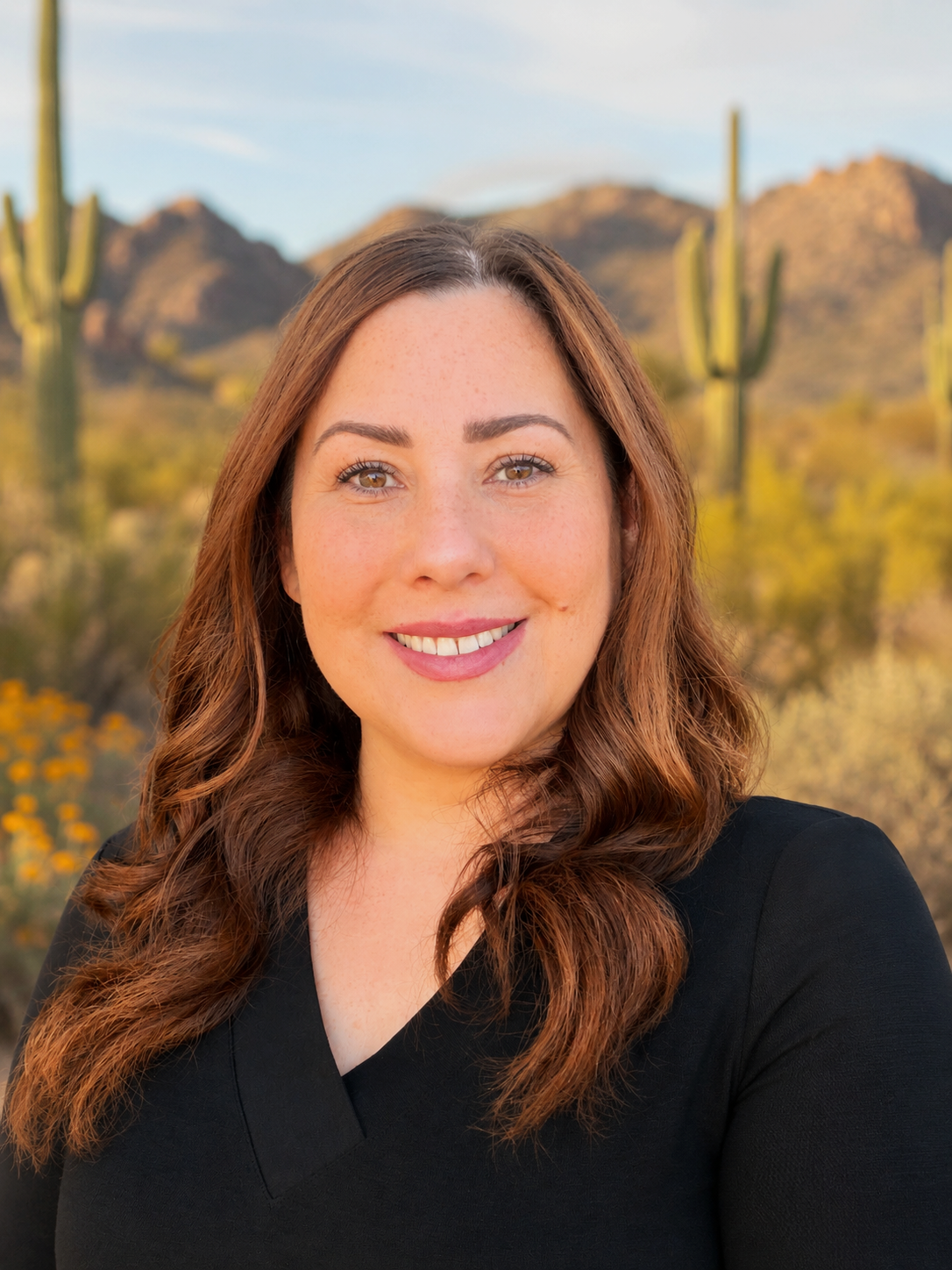 A woman with red hair, wearing large round glasses, smiling with mountains, cacti, and a sunset in the background.