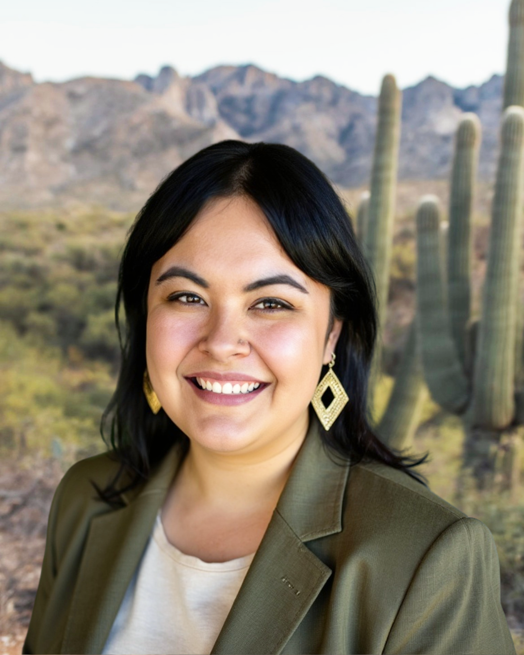 Person smiling outdoors in a desert landscape with cacti and mountains in the background.