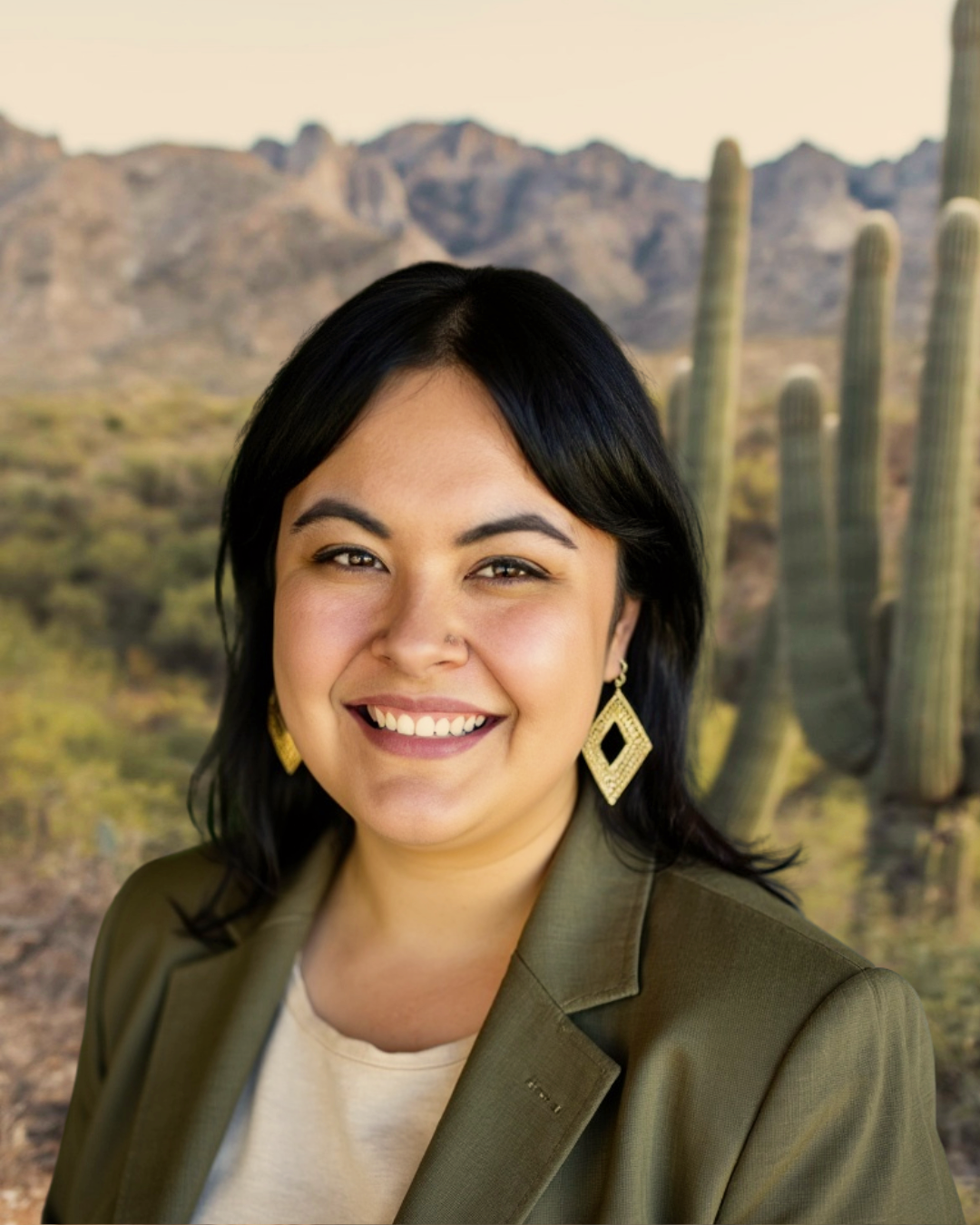Person smiling outdoors in a desert landscape with cacti and mountains in the background.
