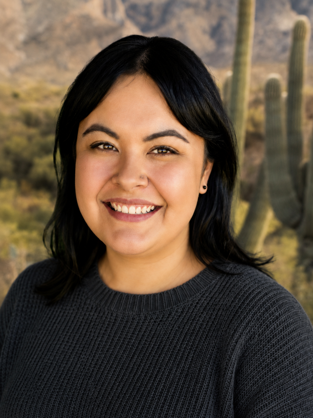 Person smiling outdoors in a desert landscape with cacti and mountains in the background.