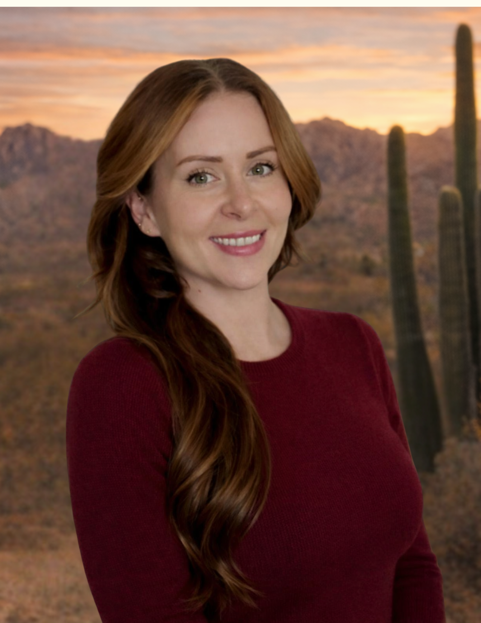 A woman with long red hair wearing a burgundy top, smiling, with a desert landscape featuring cacti and mountains at sunset in the background.