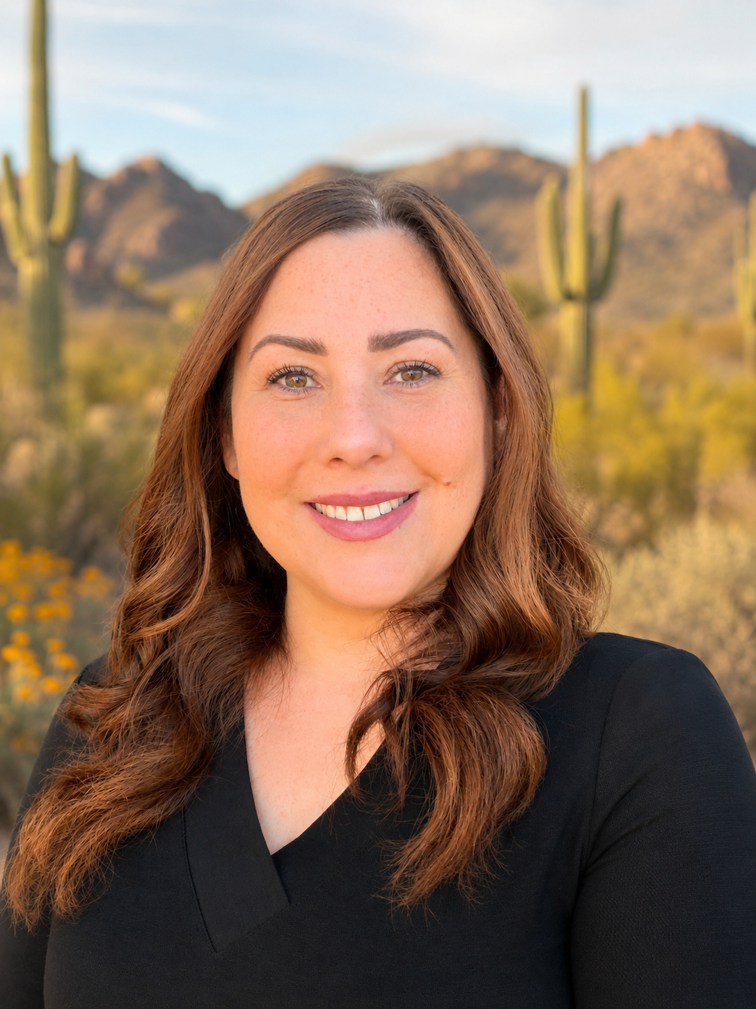 A woman with red hair, wearing large round glasses, smiling with mountains, cacti, and a sunset in the background.