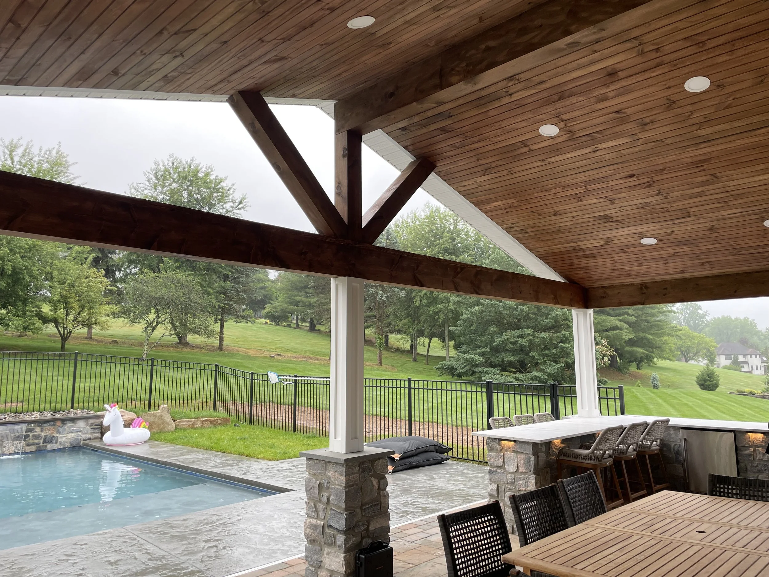Covered patio area with stone columns, wooden ceiling, and outdoor furniture. View of a backyard with a fence, grass, trees, and a swimming pool with an inflatable unicorn float.