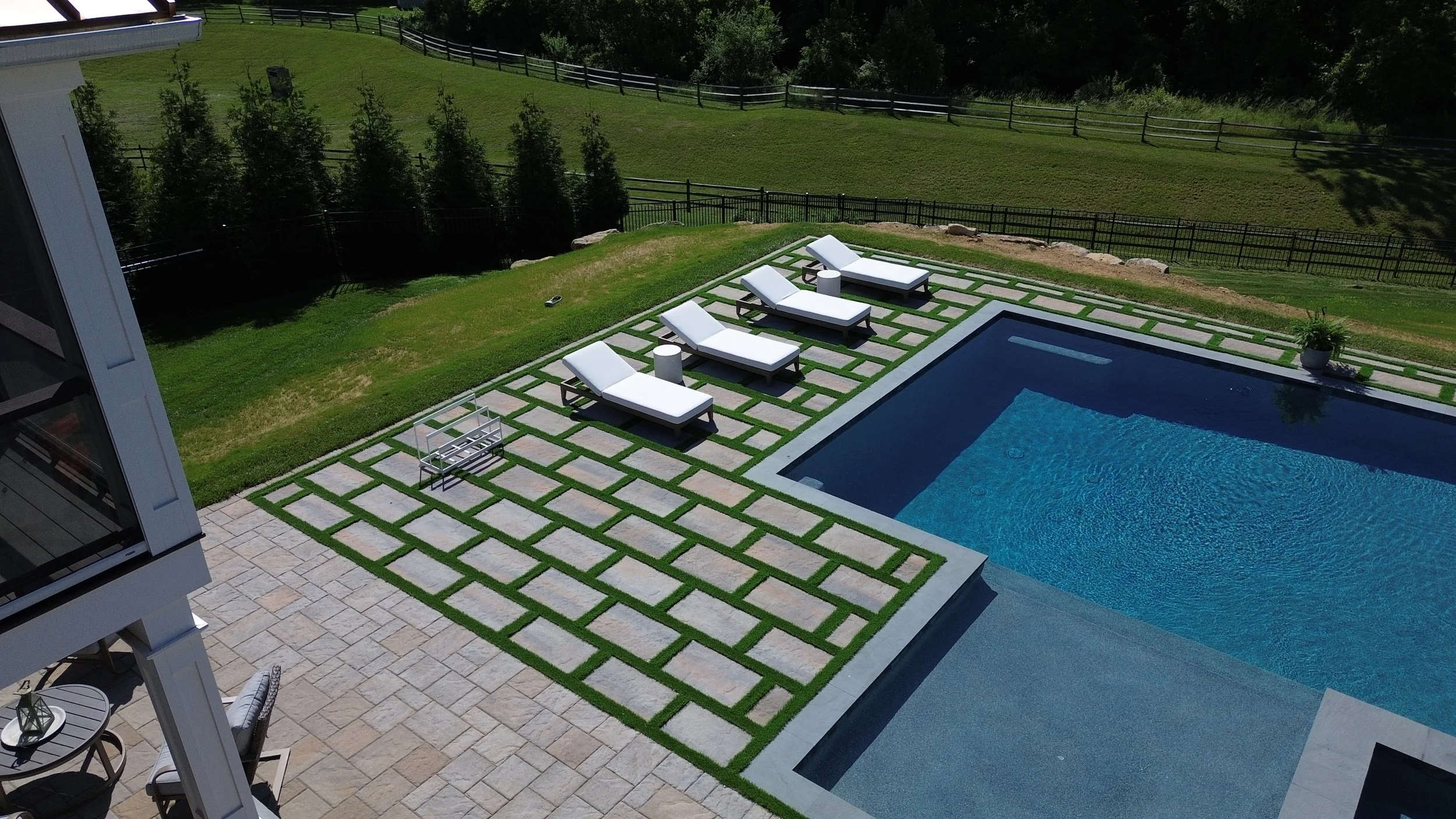 An aerial view of a backyard featuring a rectangular swimming pool with a lounge area that has four white poolside chairs and two small white tables. The area is paved with patterned tiles and has a grassy section with a small seating area. A black fence surrounds the backyard, and there are trees and greenery beyond the fence.
