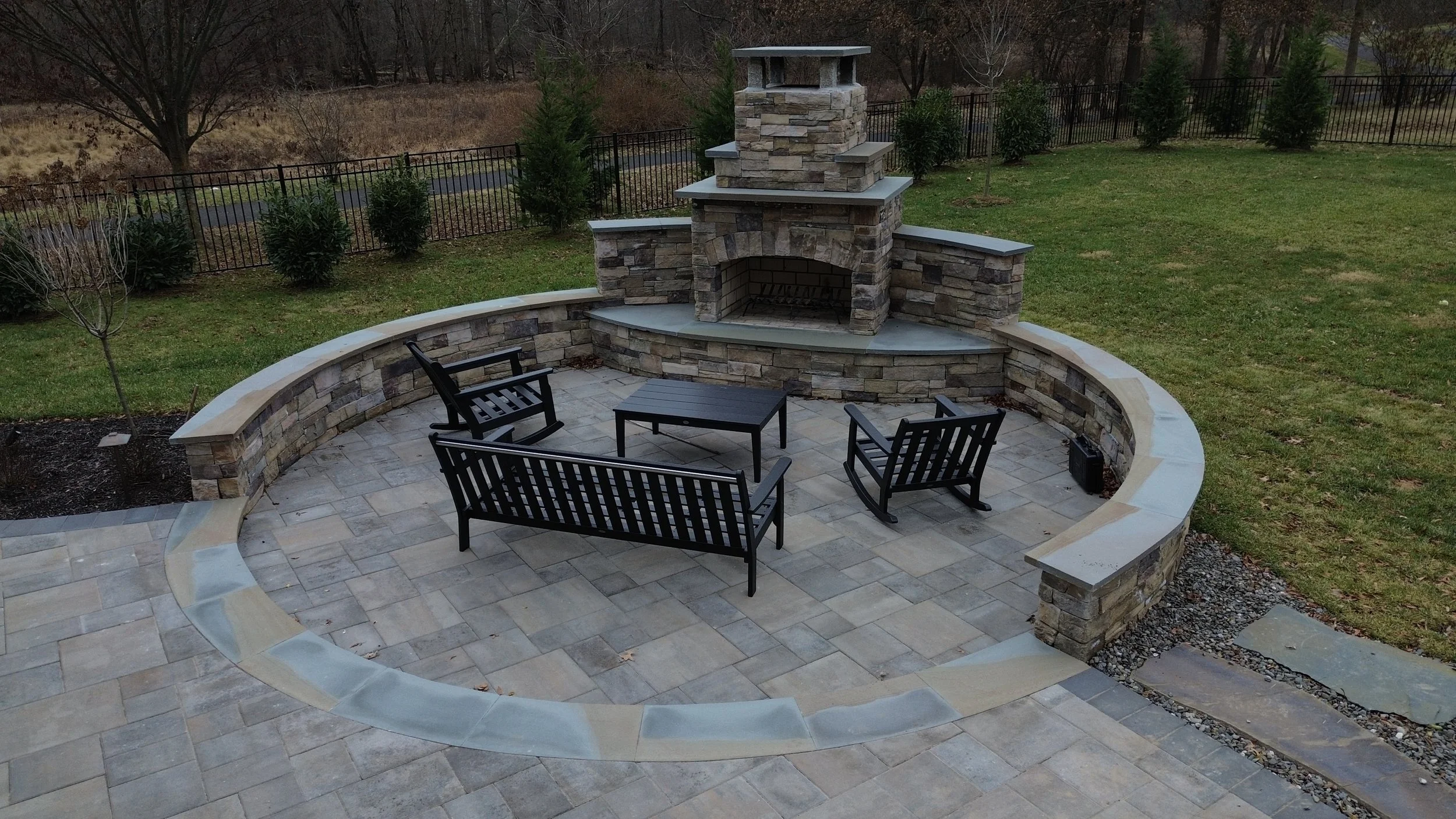 Outdoor stone fire pit area with four black patio chairs and a small black table, surrounded by a stone wall and paved patio, with a grassy yard and trees in the background.
