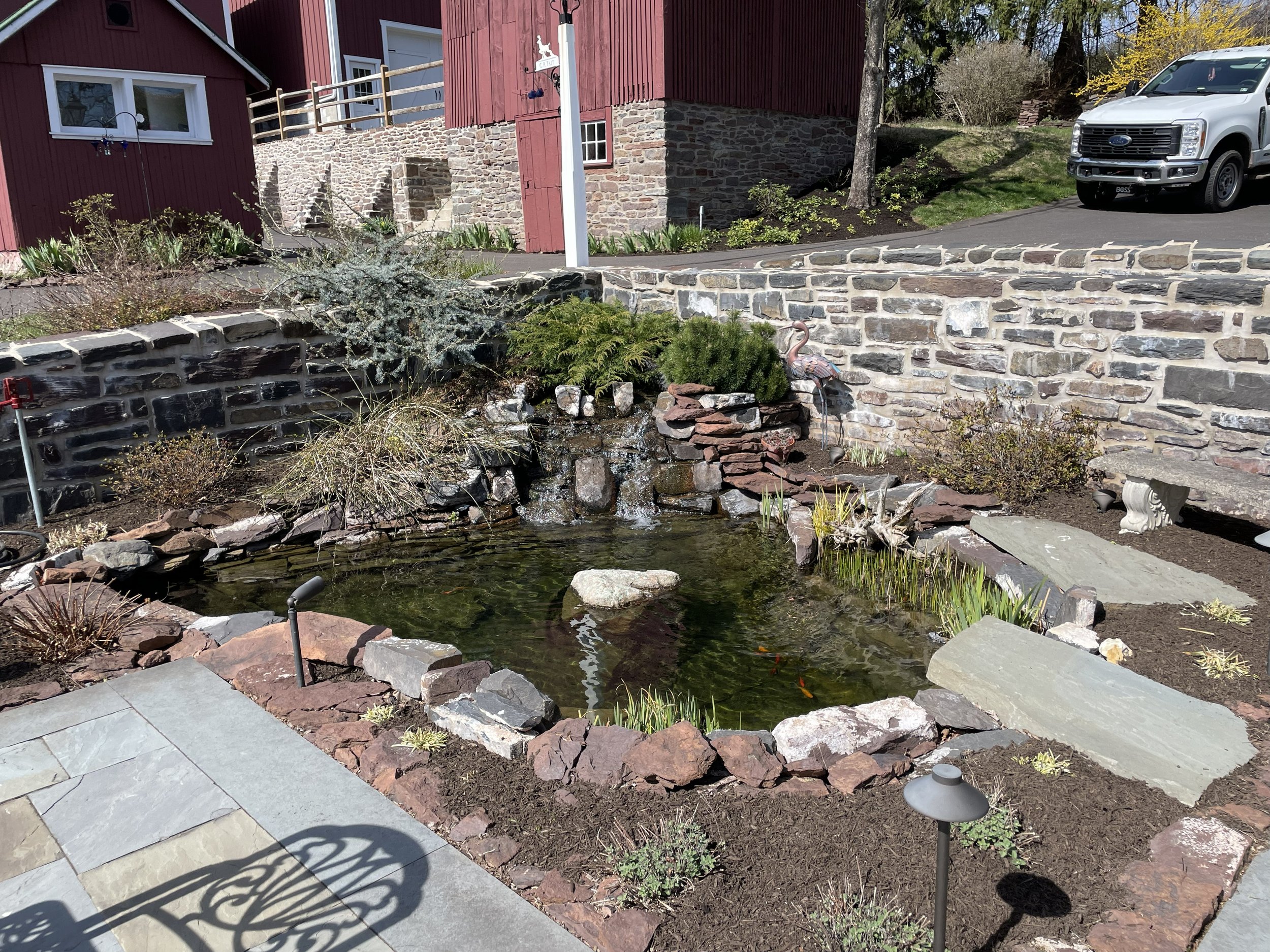 A small pond with koi fish surrounded by rocks and plants in a landscaped yard. There is a stone path and a small waterfall feature, with a building and a white truck parked nearby in the background.