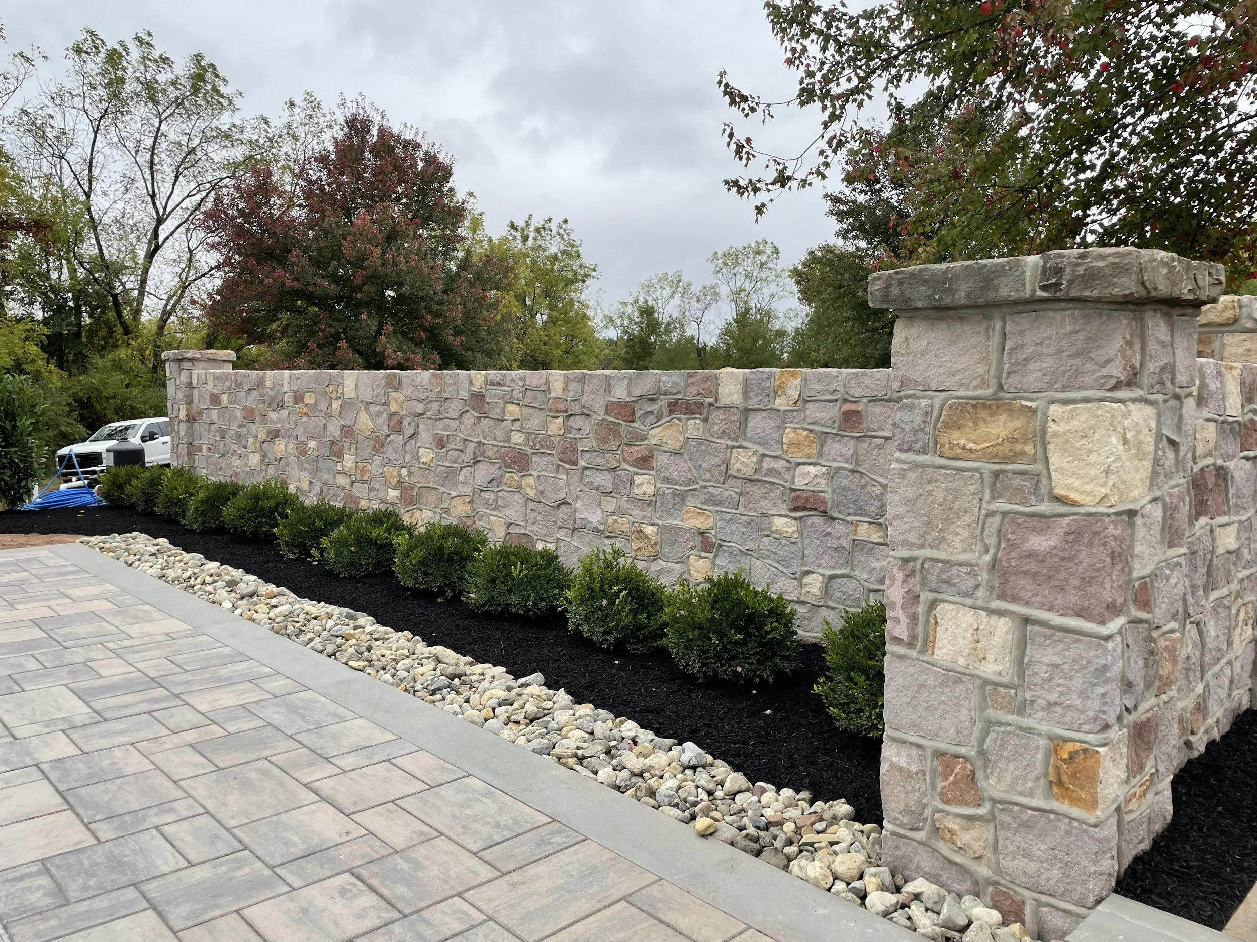 Stone wall with landscaping and bushes in front of a paved walkway, overcast sky.