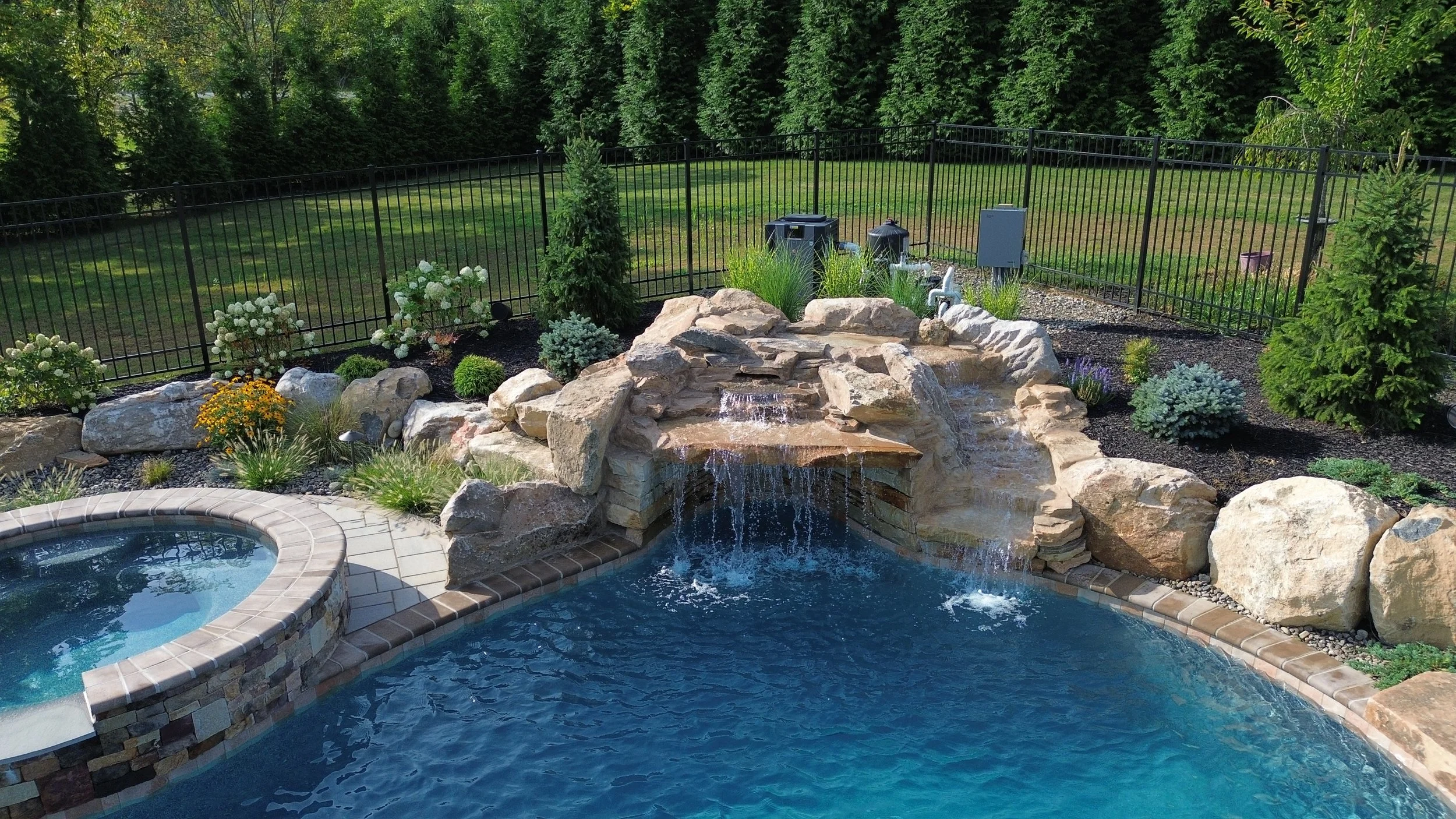 Backyard with a swimming pool, hot tub, waterfall feature surrounded by rocks and plants, fenced yard with grass and trees in the background.
