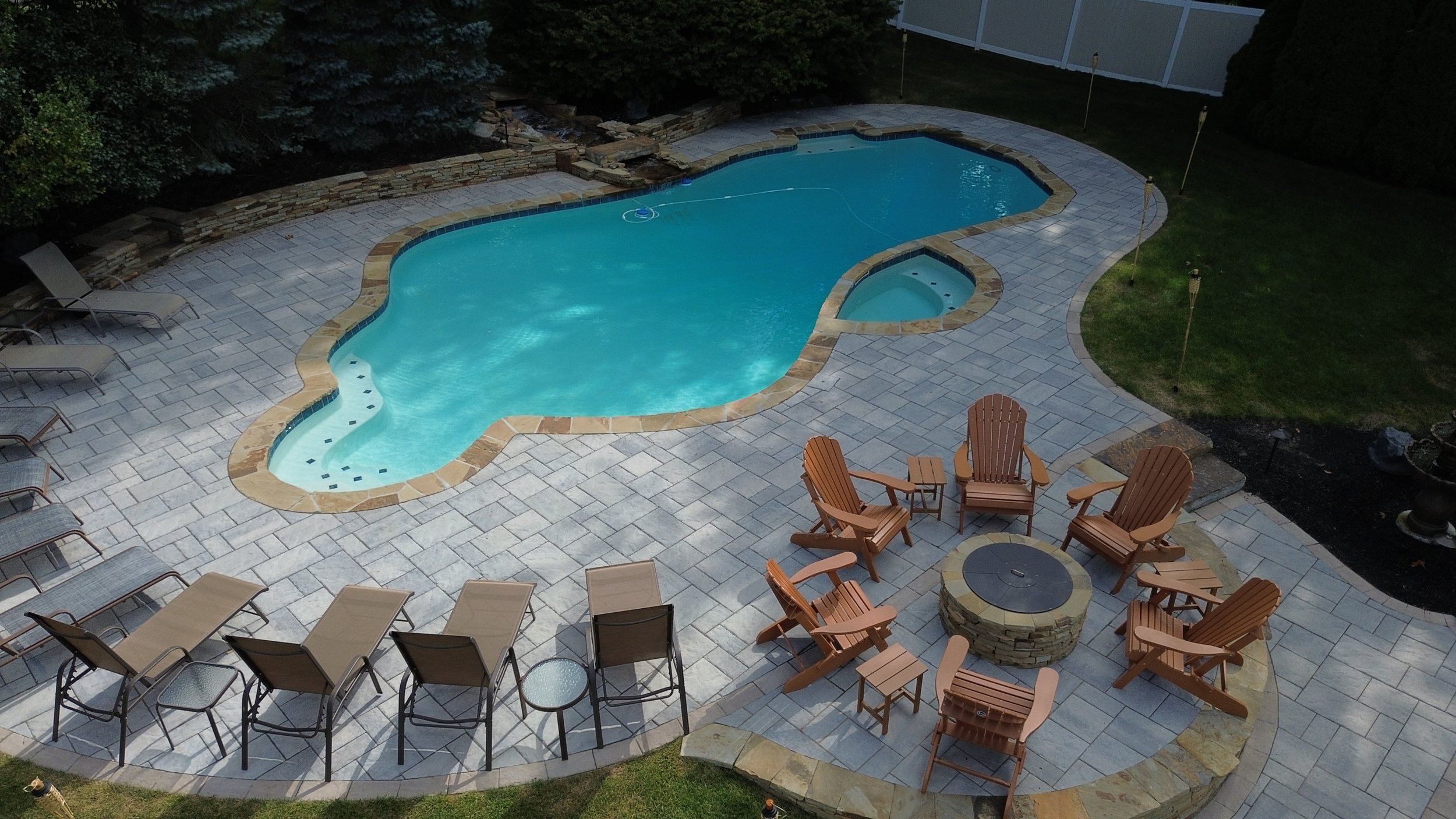 An aerial view of a backyard swimming pool with a unique shape, surrounded by stone and paver decking, with a seating area featuring wooden chairs and a fire pit, and lounge chairs along the side.