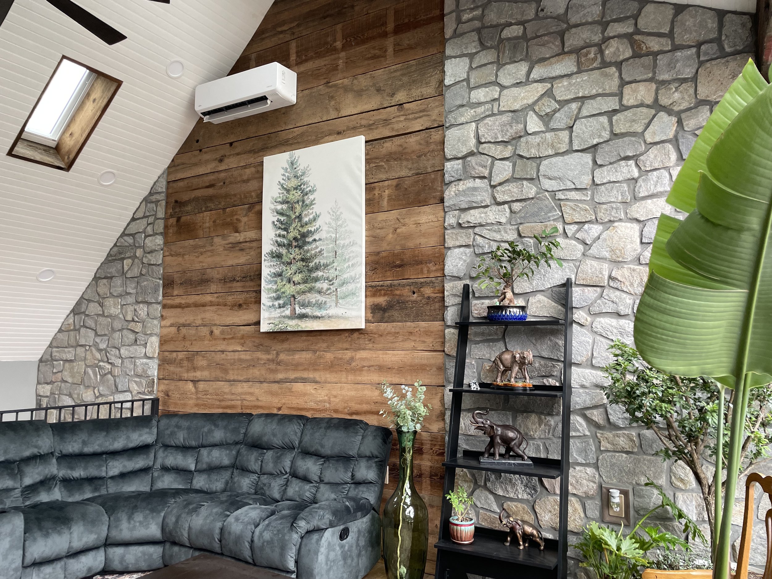 Living room with a stone and wood accent wall, black sofa, a large green vase with eucalyptus, a black shelving unit with decorative elephant and animal figurines, and potted plants; ceiling with skylight and air conditioning unit.