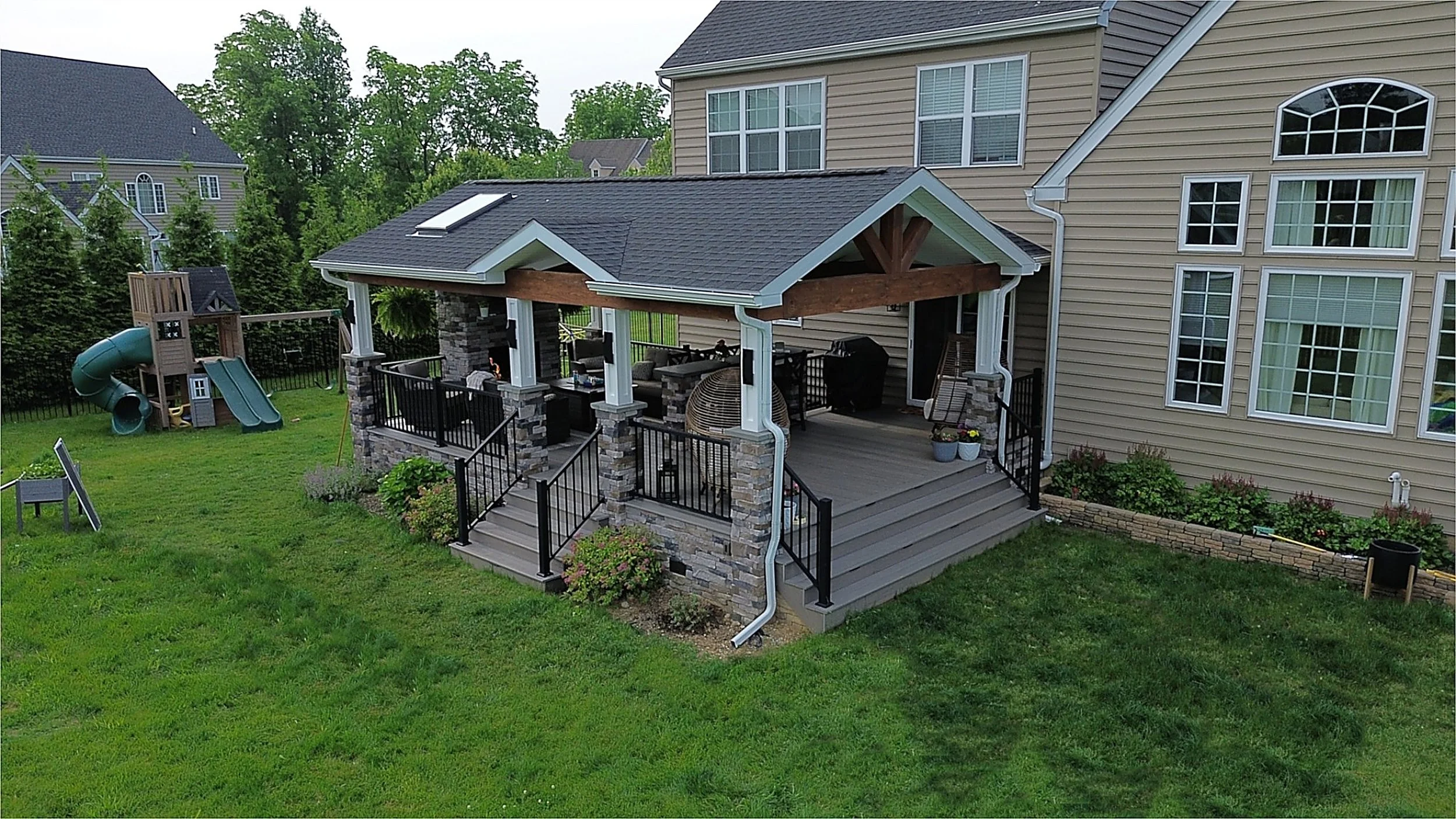 Backyard with a multi-level wooden deck with stone columns, outdoor furniture, grill, and potted plants. In the lawn, there is a children's playset with a slide and a tube slide. The yard is fenced with trees and neighboring houses in the background.