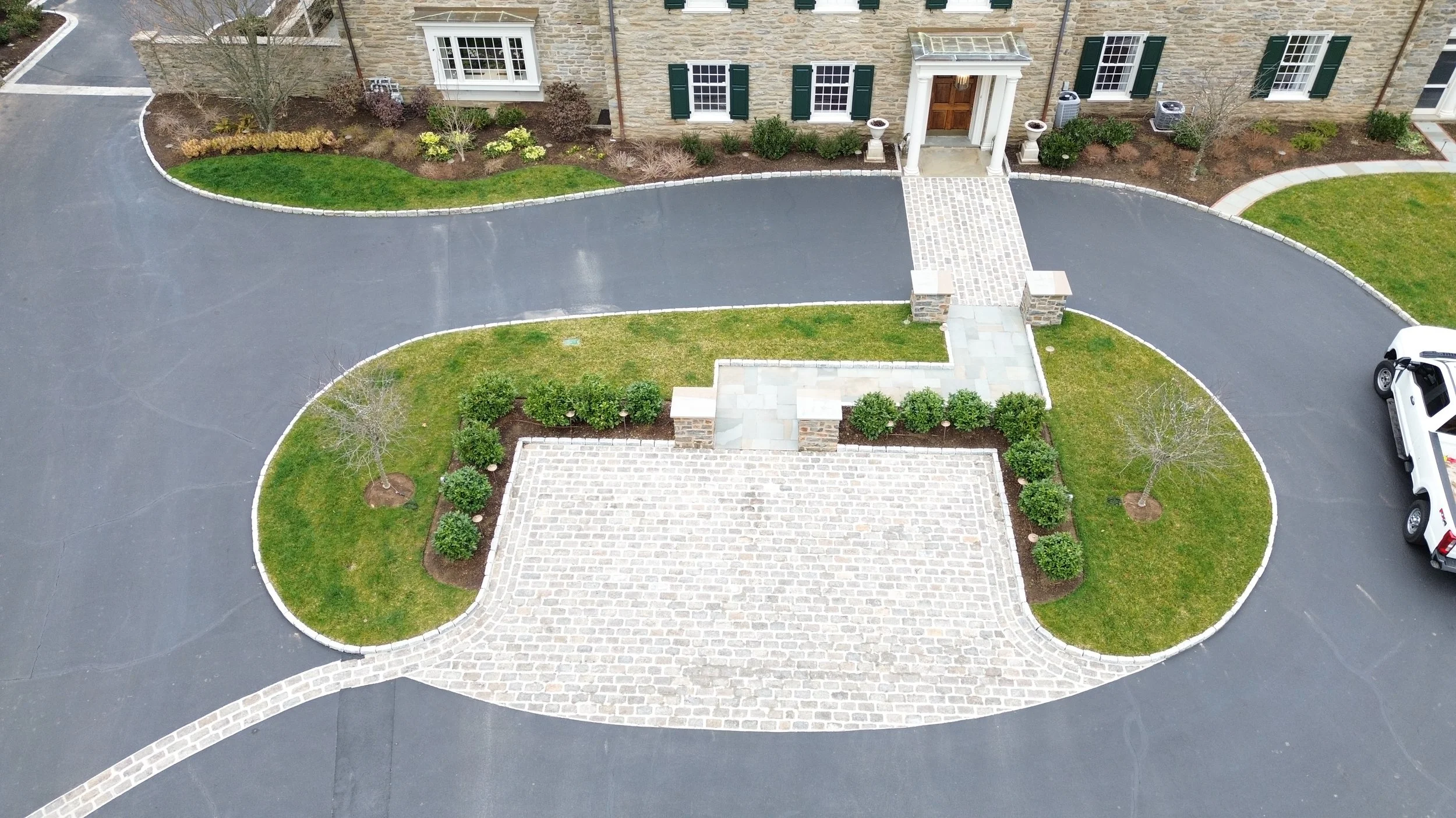Aerial view of a residential house with a curved asphalt driveway, front yard with green grass, bushes, small trees, and a paved brick walkway leading to the front door. A white vehicle is parked on the right side of the driveway.