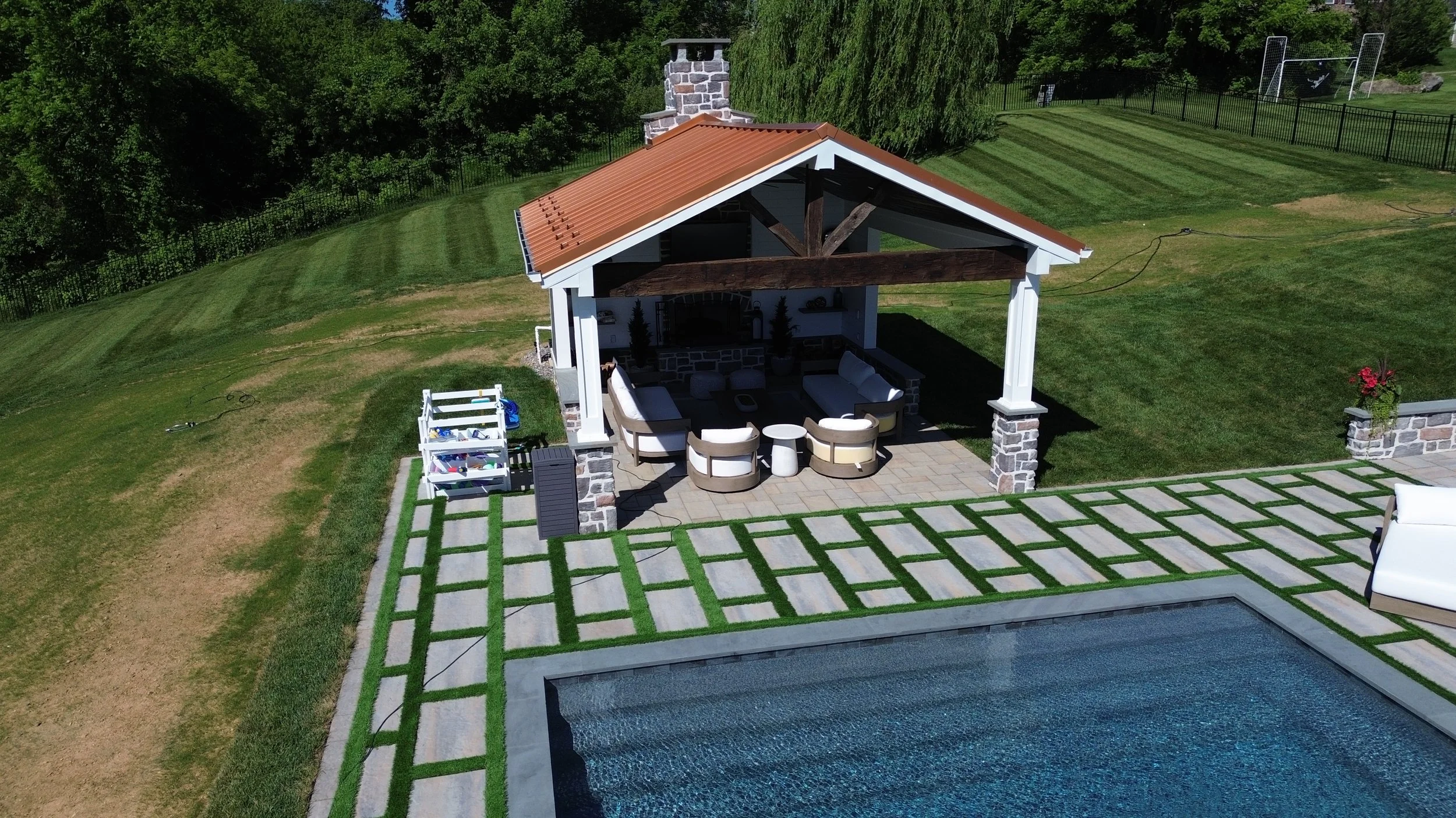 A backyard featuring a pool surrounded by a patterned concrete deck with grass in between, a covered patio area with outdoor seating and a fire pit, and a grassy lawn with trees and a sports field fence in the background.