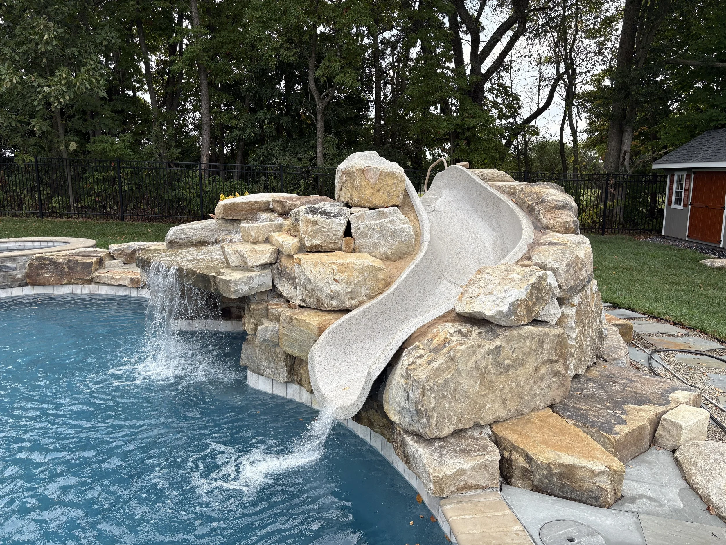 Backyard with a rock water slide leading to a swimming pool, a small shed, and trees in the background.