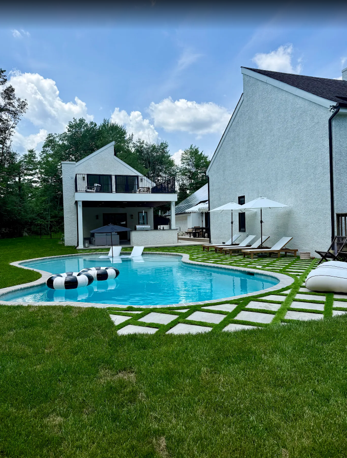 A backyard with a swimming pool surrounded by green grass and patio pavers, with lounge chairs, umbrellas, and a two-story house in the background, under a partly cloudy sky.